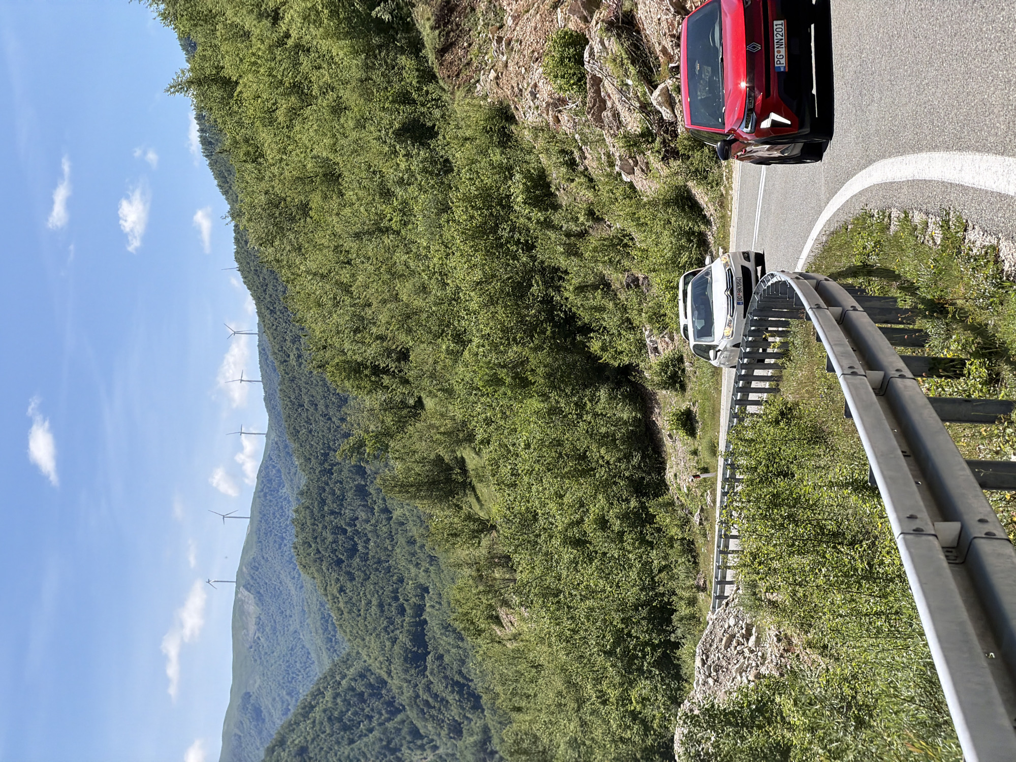 This media file presents a scenic view of a winding mountain road in Šavnik, Montenegro, captured on a clear, sunny day. The image features a two-lane asphalt road curving through a verdant, mountainous landscape.
In the foreground and midground, two cars are visible on the road. A red, modern passenger car is positioned further along the road, with its front end facing away from the viewer. Its license plate, "PG-NN201", is clearly legible. Closer to the viewer, a white passenger vehicle is situated on the right side of the road, adjacent to a metal guardrail. The white car's license plate reads "PG-BR584".
The road is bordered by dense green foliage, with steep, forested slopes rising on both sides and extending into the background. On a distant mountain ridge, several white wind turbines stand tall against the horizon, indicating a wind farm. The sky above is a bright, clear blue, dotted with scattered white clouds, suggesting pleasant weather conditions. The strong sunlight illuminates the scene, highlighting the details of the cars, road, and the rich green vegetation. The perspective offers a view along the road as it traverses the mountainous terrain.