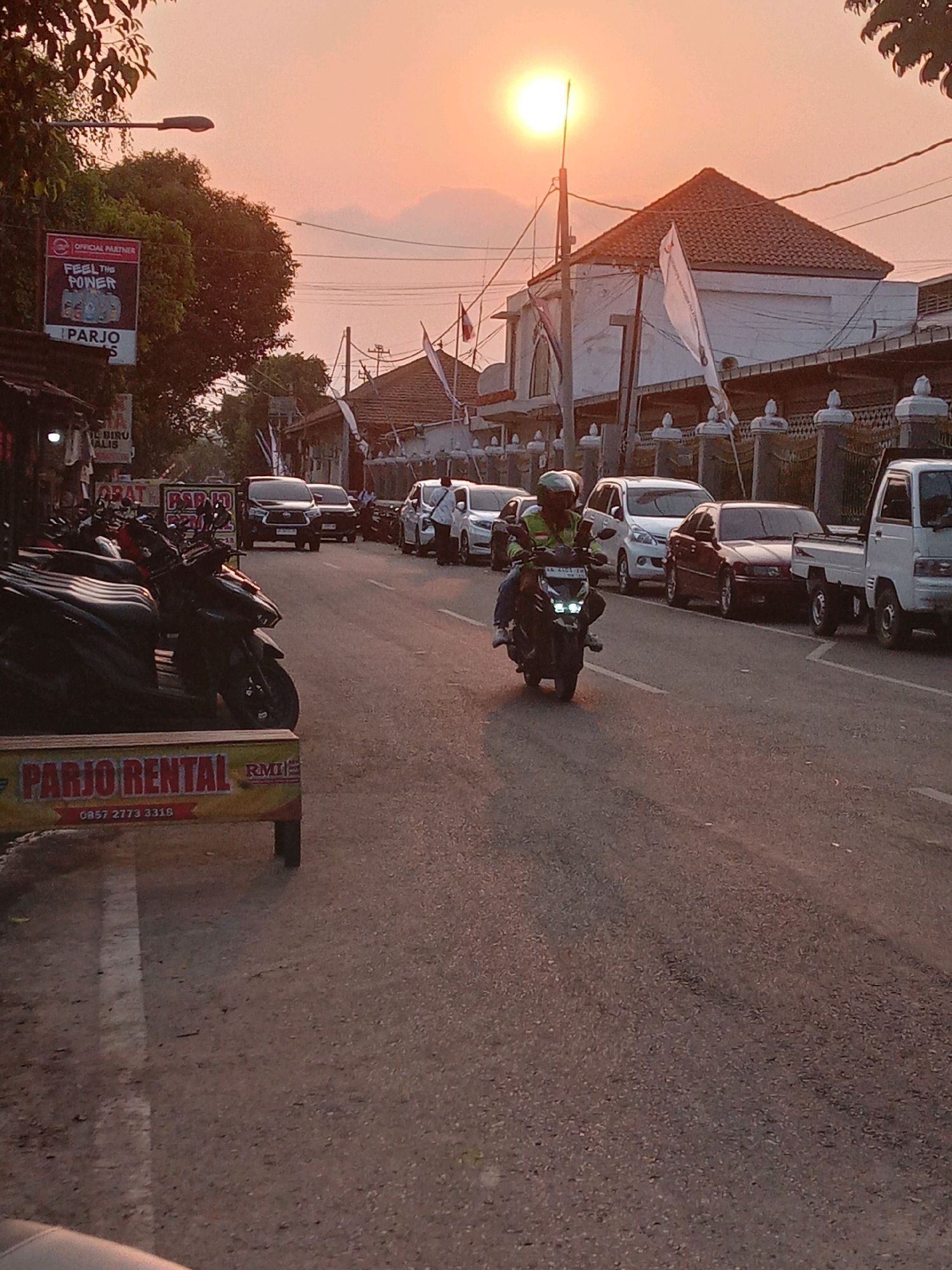 An exterior, eye-level, medium shot captures a street scene at sunset in Yogyakarta, Indonesia. The sky is a warm orange, with the sun visible as a bright circle near the top left of the frame. Silhouetted trees and power lines are visible against the sky. The street is lined with buildings, one of which has a distinctive pyramid-shaped roof. Several vehicles, including motorcycles and cars, are parked or driving on the road. In the foreground, a yellow sign with red lettering reads "PARJO RENTAL" and a phone number. A person on a motorcycle, wearing a green vest and helmet, is in the center of the frame, moving down the street. The overall impression is of a busy urban environment during a colorful sunset.