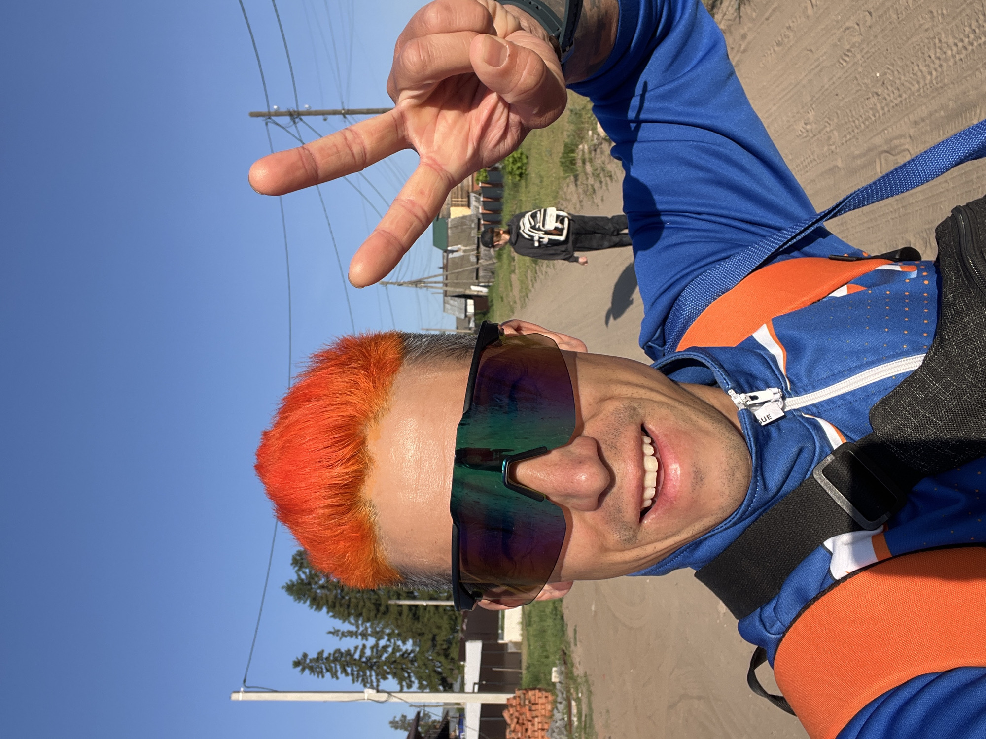 A man with bright orange hair and sunglasses is holding up a peace sign in a selfie outdoors. He is wearing a blue and white athletic jacket and an orange harness. In the background, a person in a black and white jacket is walking away on a dirt road. A dirt road is visible, with wooden buildings and power lines in the background. The photo was taken in Chaikovsky, Russia.