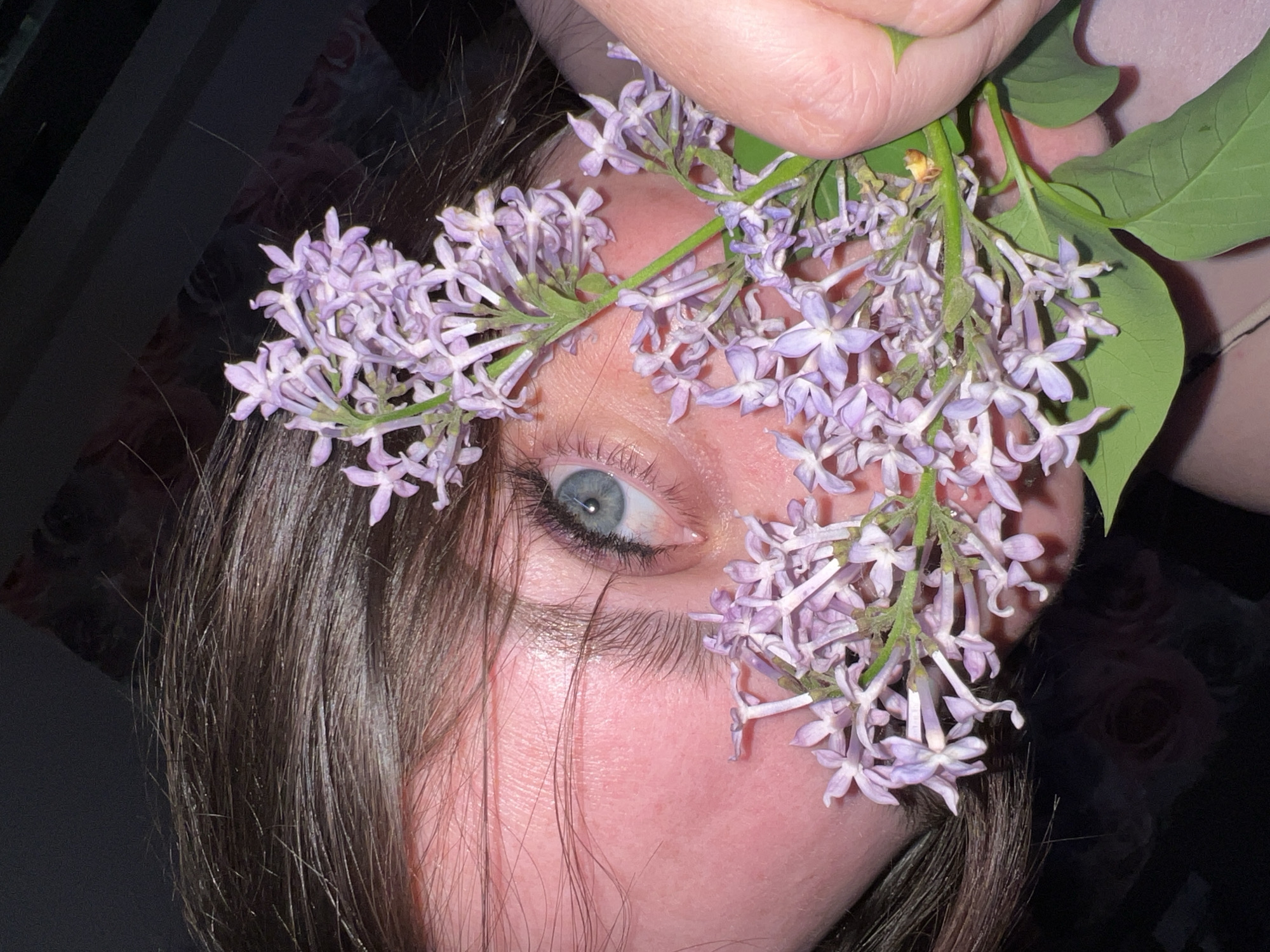 An extreme close-up photograph features a person's face obscured by lilac flowers. The individual has an eye with a light-blue iris. The person is holding several lilac flowers. The background is mostly dark. The location in the city of Noviy, Russia is not discernible from the photograph.