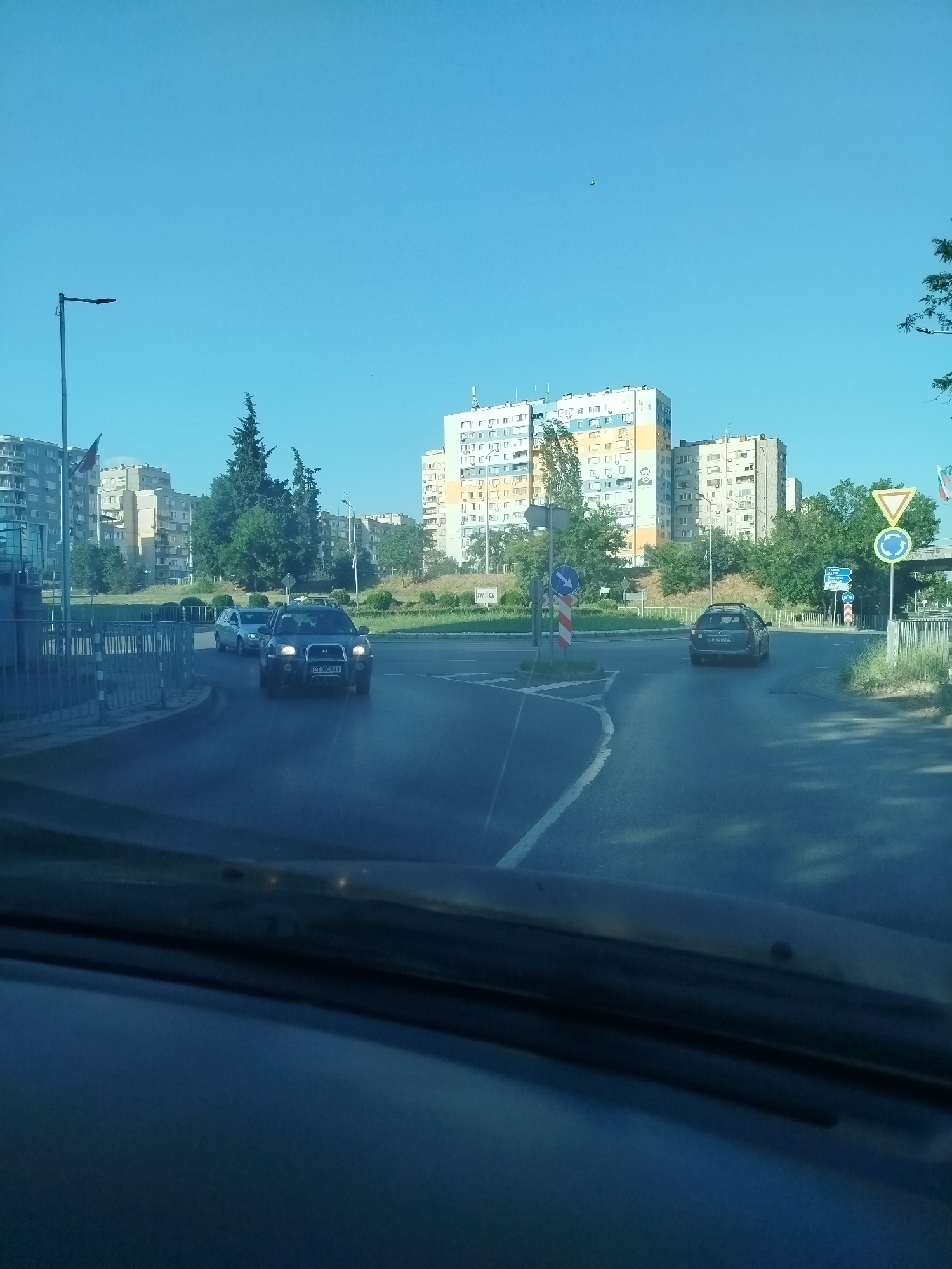The image shows a clear, blue sky over a city street. In the foreground, the dashboard and windshield of a vehicle are visible. The vehicle is moving through an intersection. Several cars are present on the road, including a dark-colored SUV in the left lane and a lighter-colored sedan in the right lane. Behind the cars, tall, multi-story apartment buildings dominate the skyline. Trees and vegetation are scattered throughout the scene. Traffic signs, including a yield sign and a roundabout sign, are visible near the intersection. The overall impression is of a typical urban environment on a bright, sunny day.