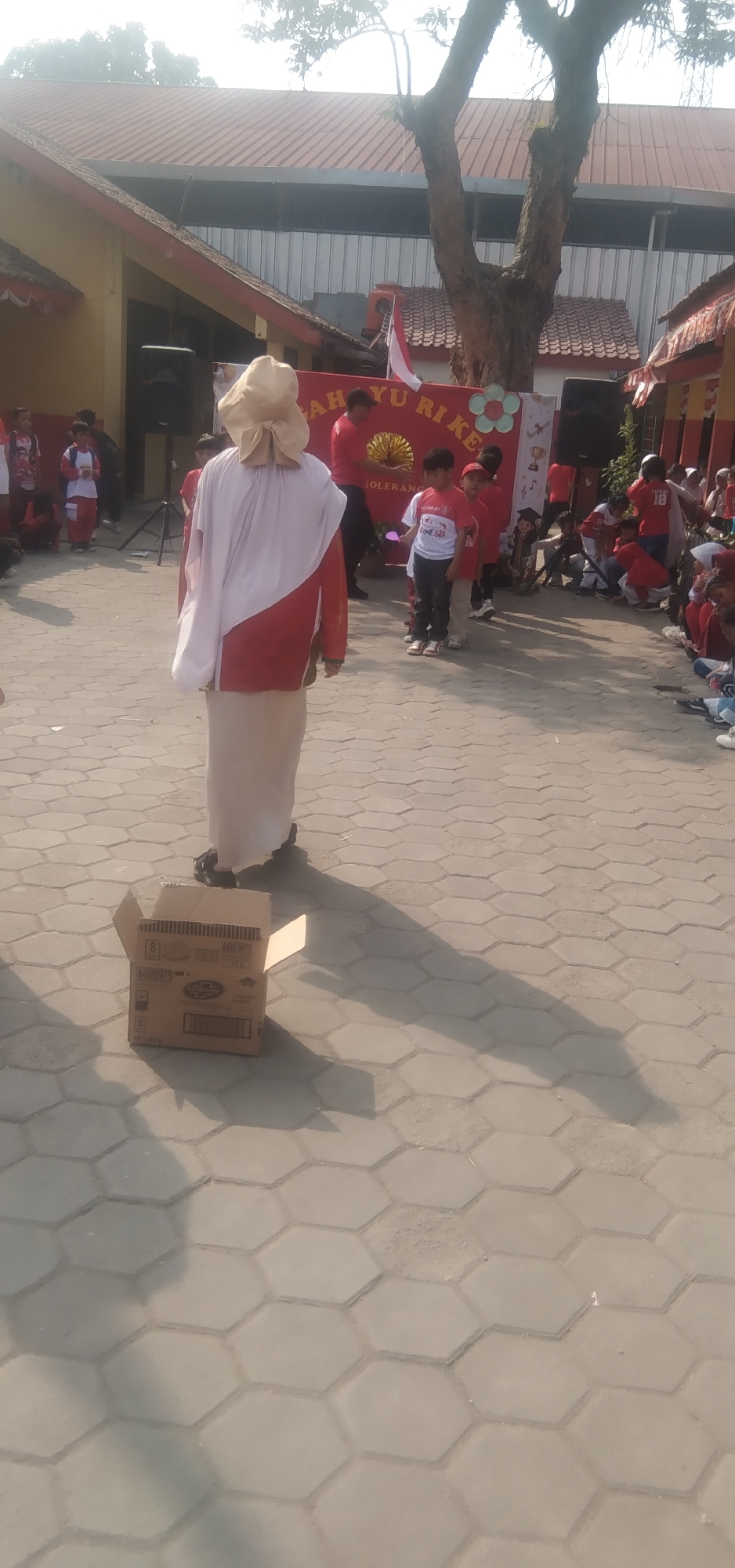 The image depicts an outdoor scene, likely in Rahayu, Indonesia, on a paved courtyard with a hexagonal tile pattern. The background features buildings with red roofs and yellow walls, typical of Indonesian architecture. In the middle ground, a group of children and adults are gathered, many wearing red and white attire, possibly participating in a local festival or event. A banner with Indonesian text is visible, indicating a celebration or announcement. In the foreground, a person in traditional Indonesian clothing, with a wide-brimmed hat, stands facing away from the camera, casting a long shadow. An open cardboard box rests on the pavement near the person. The overall atmosphere suggests a community gathering in a warm, sunny climate.
