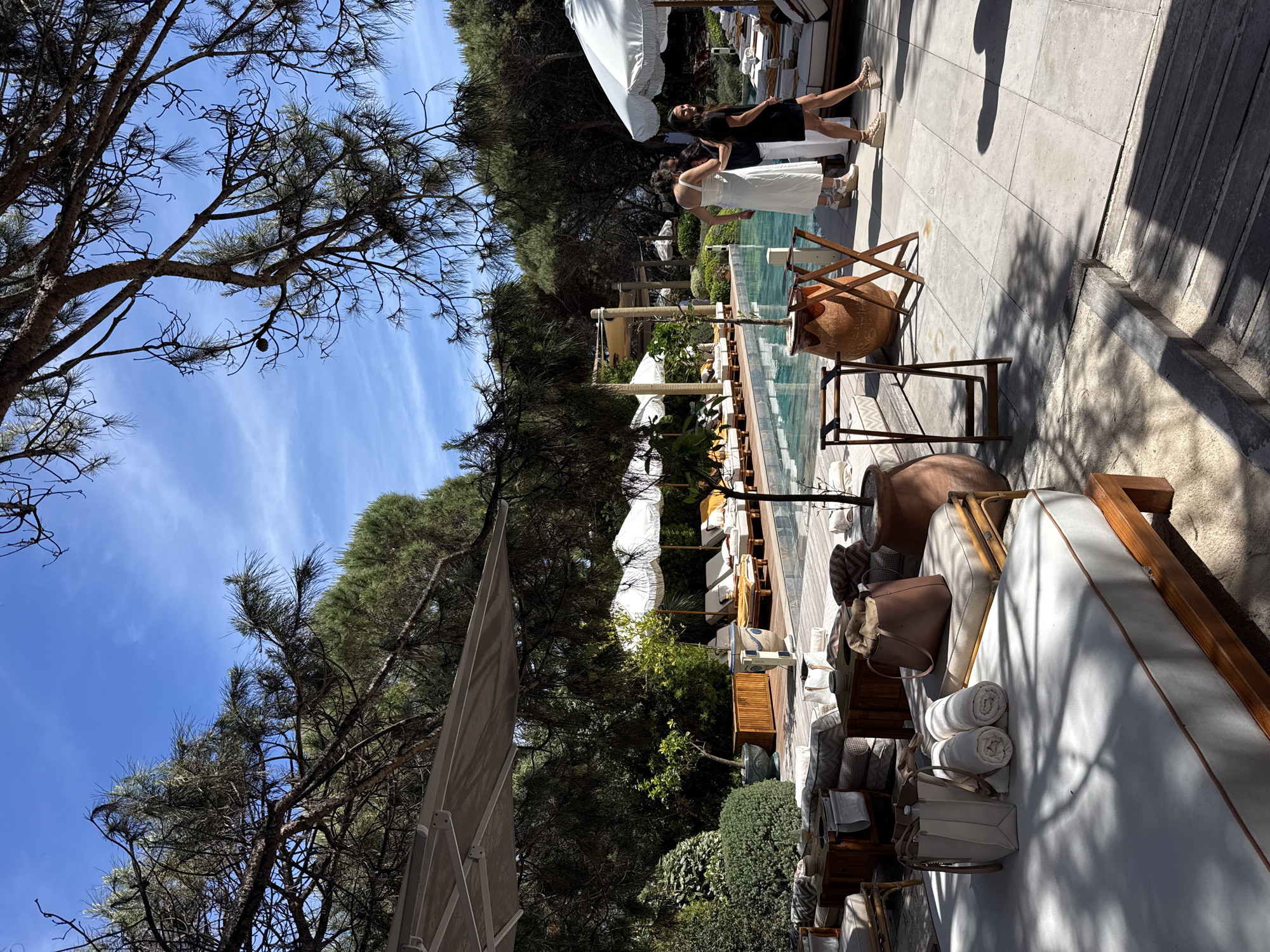 The image depicts a scene by a swimming pool in Ramatuelle, France. Two women are seen standing near the pool edge. One woman is wearing a white dress and the other is in a black dress. The pool is lined with lounge chairs under white umbrellas. The pool is surrounded by trees. There are large clay pots and bags near the lounge chairs. The image is shot during the daytime under a blue sky.
