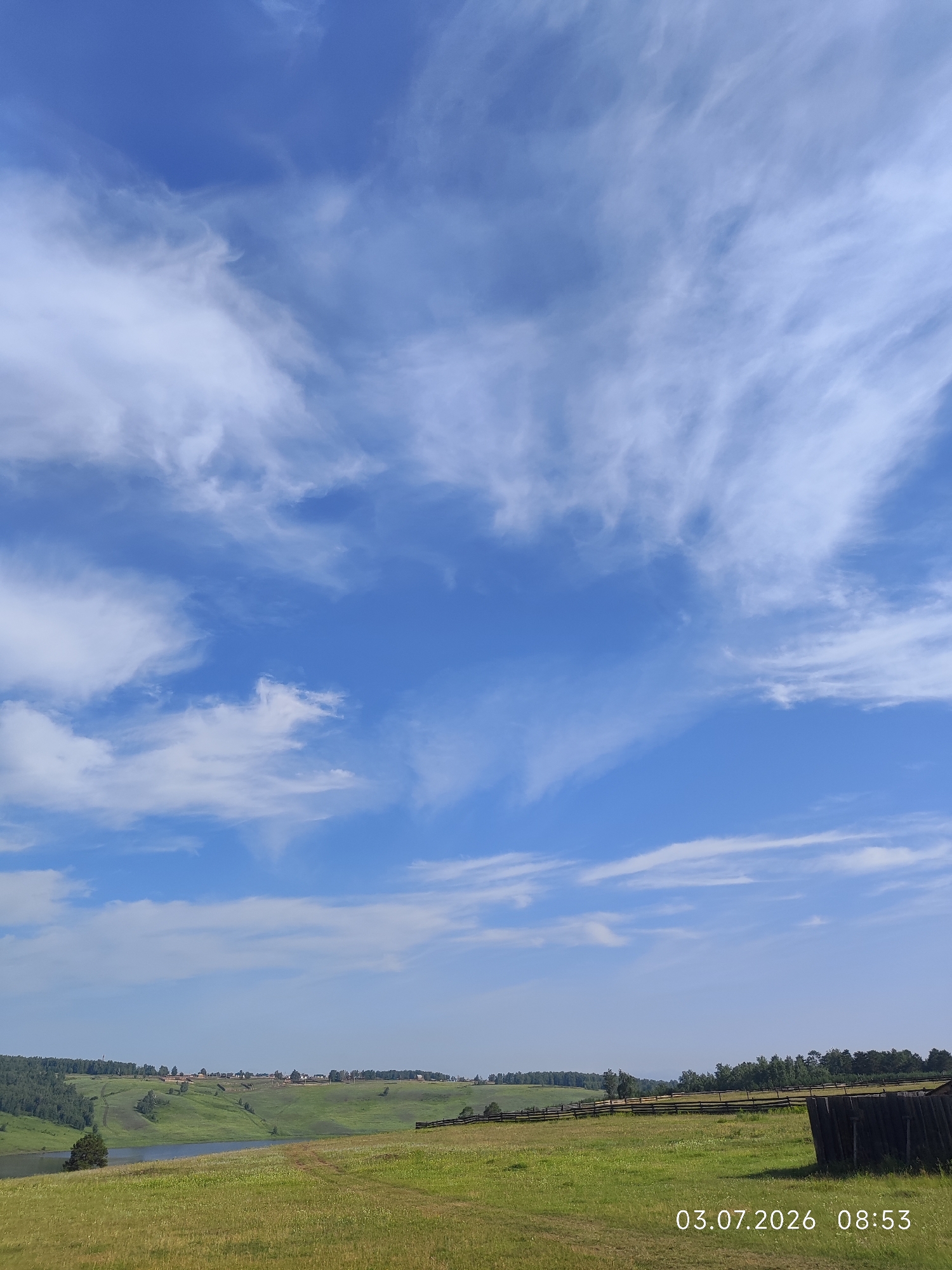 The image depicts a rural landscape with a wide expanse of blue sky filled with wispy white clouds. In the foreground, a grassy field stretches out, with faint tracks suggesting pathways. A dark wooden structure is visible on the right side of the field. Beyond the field, rolling green hills dotted with trees and small buildings form the horizon. A body of water, possibly a lake or river, is nestled among the hills. The timestamp "03.07.2026 08:53" is overlaid on the bottom right of the image, indicating the date and time it was taken. The scene appears to be a peaceful, natural environment in Konovalovo, Russia.