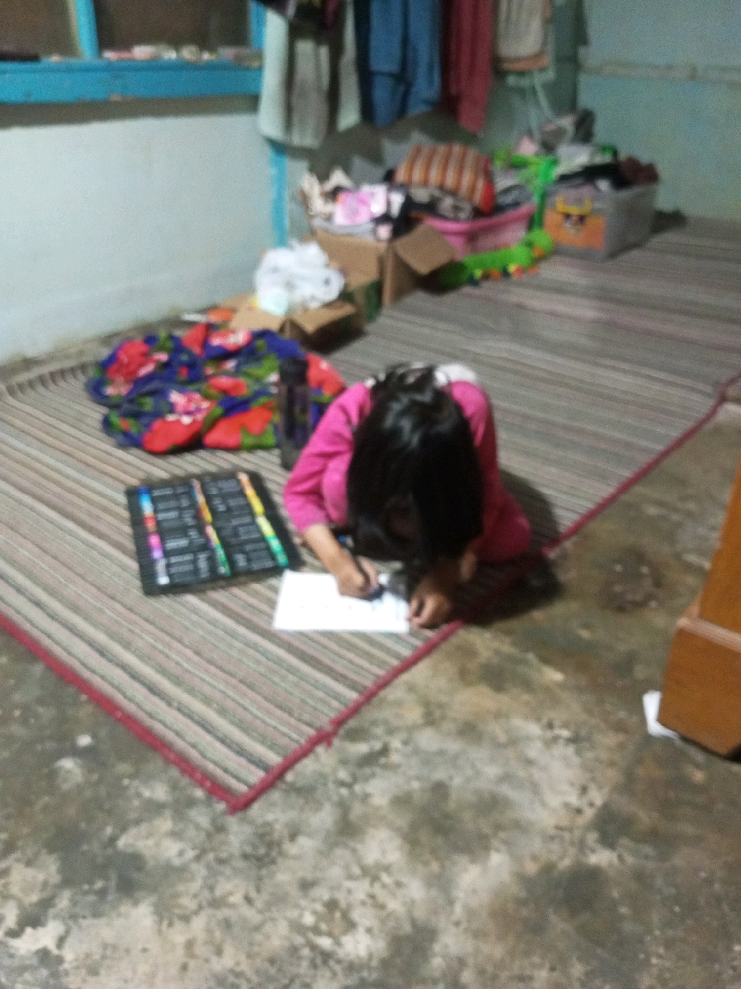 In Andongsili, Indonesia, a child kneels on a woven mat, engrossed in drawing. The child wears a bright pink long-sleeved shirt and has dark hair. In front of the child lies a box of colored markers, neatly arranged. Adjacent to the child, a colorful blanket is spread out, featuring a floral pattern of red and blue. The room appears somewhat cluttered, with various items visible in the background, including boxes, clothing hanging, and a plastic bin. The floor is a rough concrete surface.