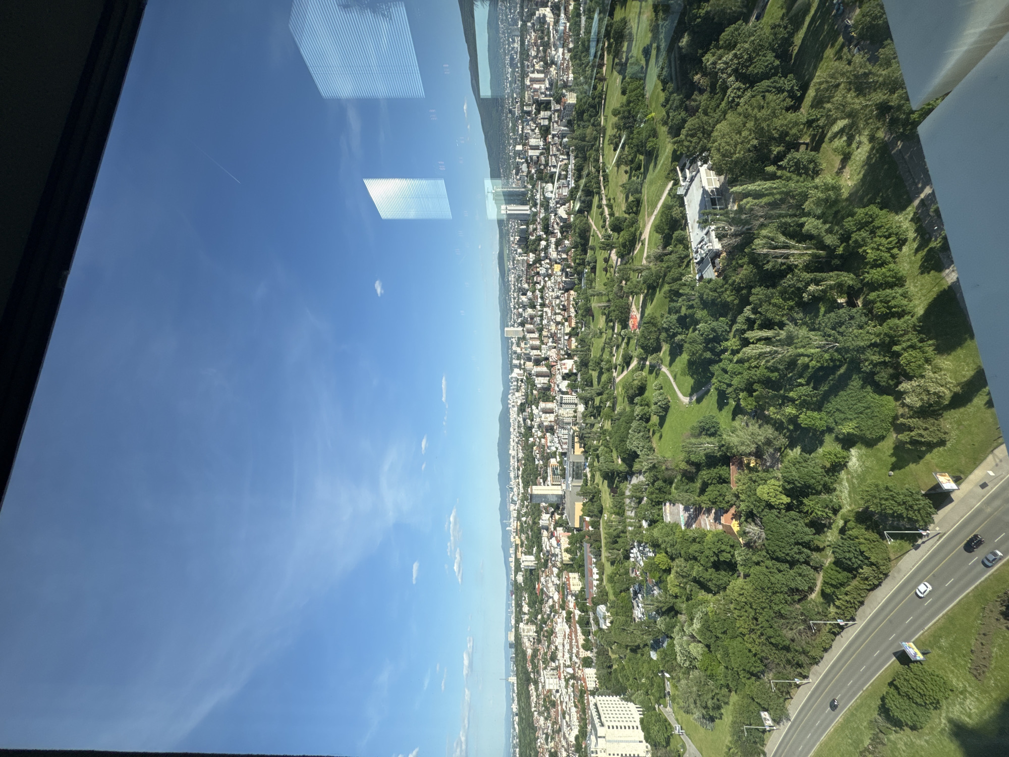 The image presents an elevated view of Sofia, Bulgaria, likely taken from a high-rise building. The foreground features a window frame, with reflections of interior blinds and sky visible on the glass. The city is divided into two main sections. One section includes a dense urban area with numerous buildings of varying heights, suggesting a mix of residential and commercial zones. A mountainous region is visible in the distance. The other section features a large park with extensive green spaces, trees, and winding paths. A main road runs along the edge of the park with visible cars.