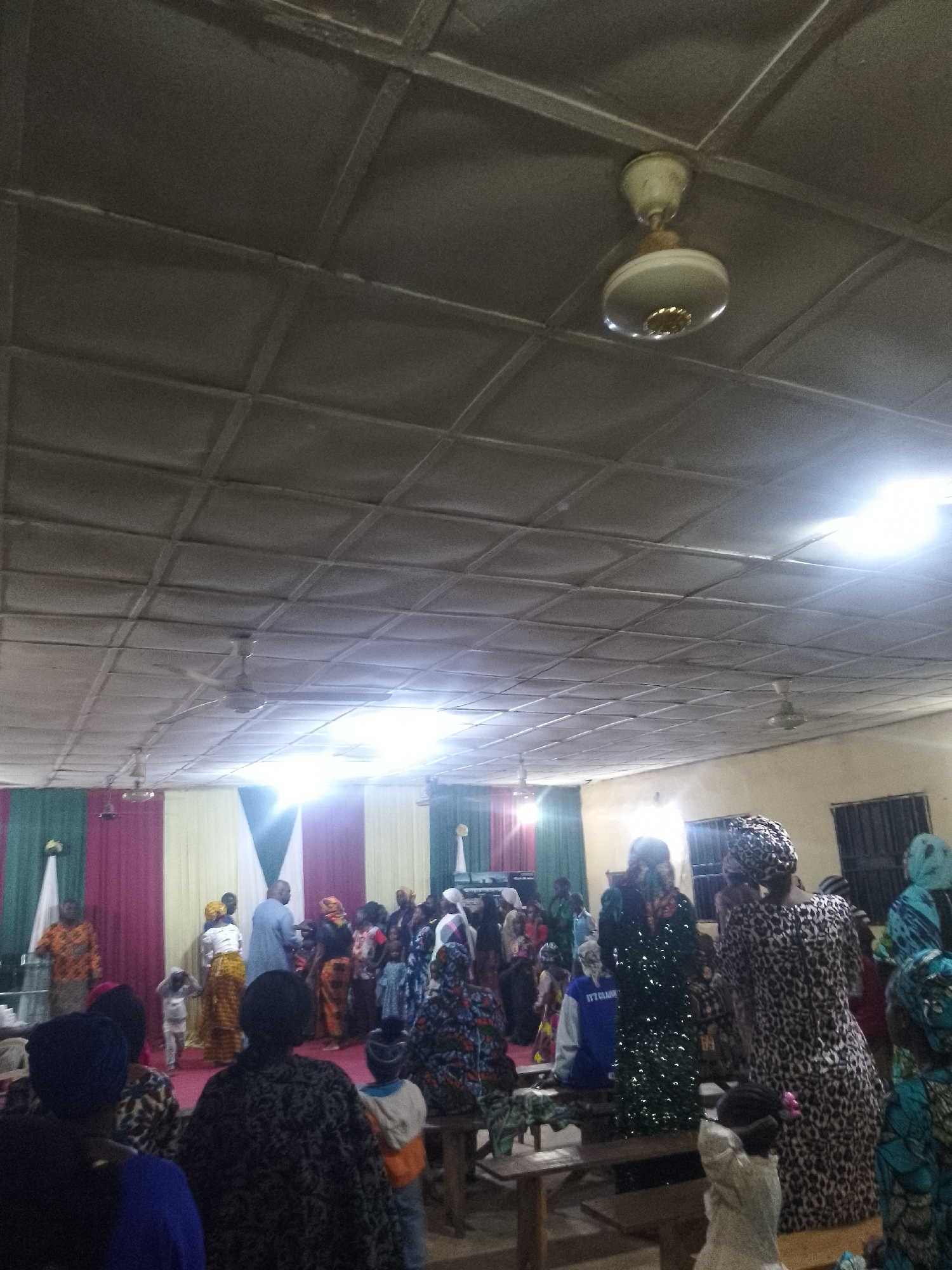 The image depicts an indoor gathering, likely a religious service or celebration, in Serti, Nigeria. A group of people, including men, women, and children, are present in a room with a textured, grid-like ceiling. Several ceiling fans are visible, suggesting a warm climate. The room is decorated with draped fabric in shades of green, pink, and cream, forming a backdrop that appears to be a stage or focal point of the gathering.
In the foreground, several individuals are facing away from the camera, their backs visible. They are dressed in various patterned fabrics, typical of Nigerian attire. Some are wearing headwraps. In the mid-ground and background, more people are engaged in activities, with some appearing to be standing and conversing, while others might be seated or observing. One person in the background is wearing a blue garment. The lighting is bright, with some glare suggesting artificial lights overhead. The overall atmosphere appears to be one of community and shared occasion.