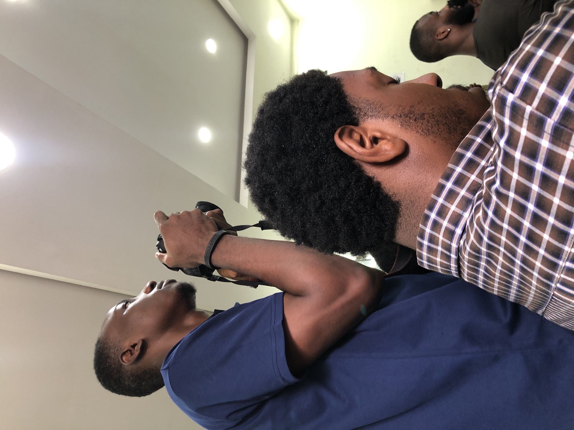 In Ikeja, Nigeria, two men are captured in a medium shot, with one in the foreground and another slightly behind and to the left. The man in the foreground, facing right and looking downwards, has dark, curly hair and is wearing a brown and white checkered shirt. His profile is visible, showcasing his ear and part of his beard. Behind him and to the left, a man wearing a blue t-shirt is holding a camera to his face, appearing to take a photograph. His head is tilted upwards, and he is looking towards something out of frame. A black wristband with white text is visible on his wrist. The background consists of a modern ceiling with recessed lighting, suggesting an indoor setting. The lighting is bright, illuminating the scene.