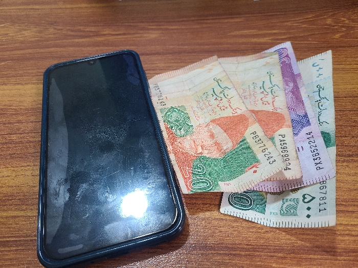 The scene is located indoors in Quetta City Tehsil, Pakistan. A dark-colored smartphone, enclosed in a dark blue or black protective case, rests horizontally on a reddish-brown wooden surface with visible grain. The smartphone's screen is off, displaying smudges, minor scratches, and a circular reflection of an overhead light source. To the right of the smartphone, four Pakistani Rupee banknotes are fanned out and partially overlapping. From top to bottom, these include two 20 Rupee notes (PKR 20) with visible serial numbers PB3776243 and PA5969924, one purple 50 Rupee note (PKR 50) with serial number PX3652214, and one green and white 50 Rupee note (PKR 50) with visible serial number 867811. No individuals are present in the image.