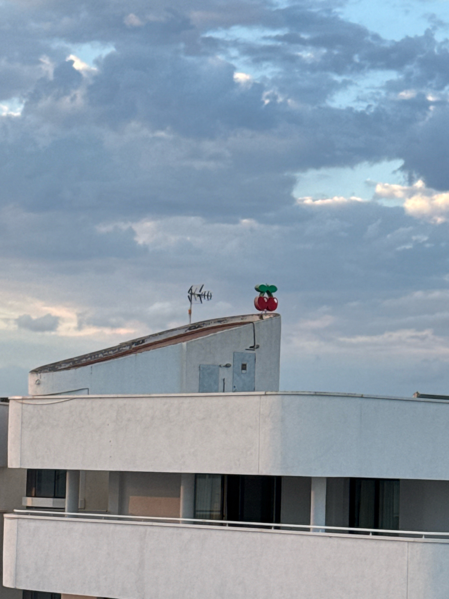 The image captures the rooftop of a building in Ibiza, Spain, under a cloudy sky. Prominently featured on the peak of the building's sloped roof is a red cherry-shaped decorative item with a green stem and leaves. To the left of the cherry decoration, an antenna is visible. The building itself is white and appears to have multiple levels with balconies and windows. The sky is filled with gray and blue clouds, suggesting overcast weather, with hints of lighter sky peeking through.