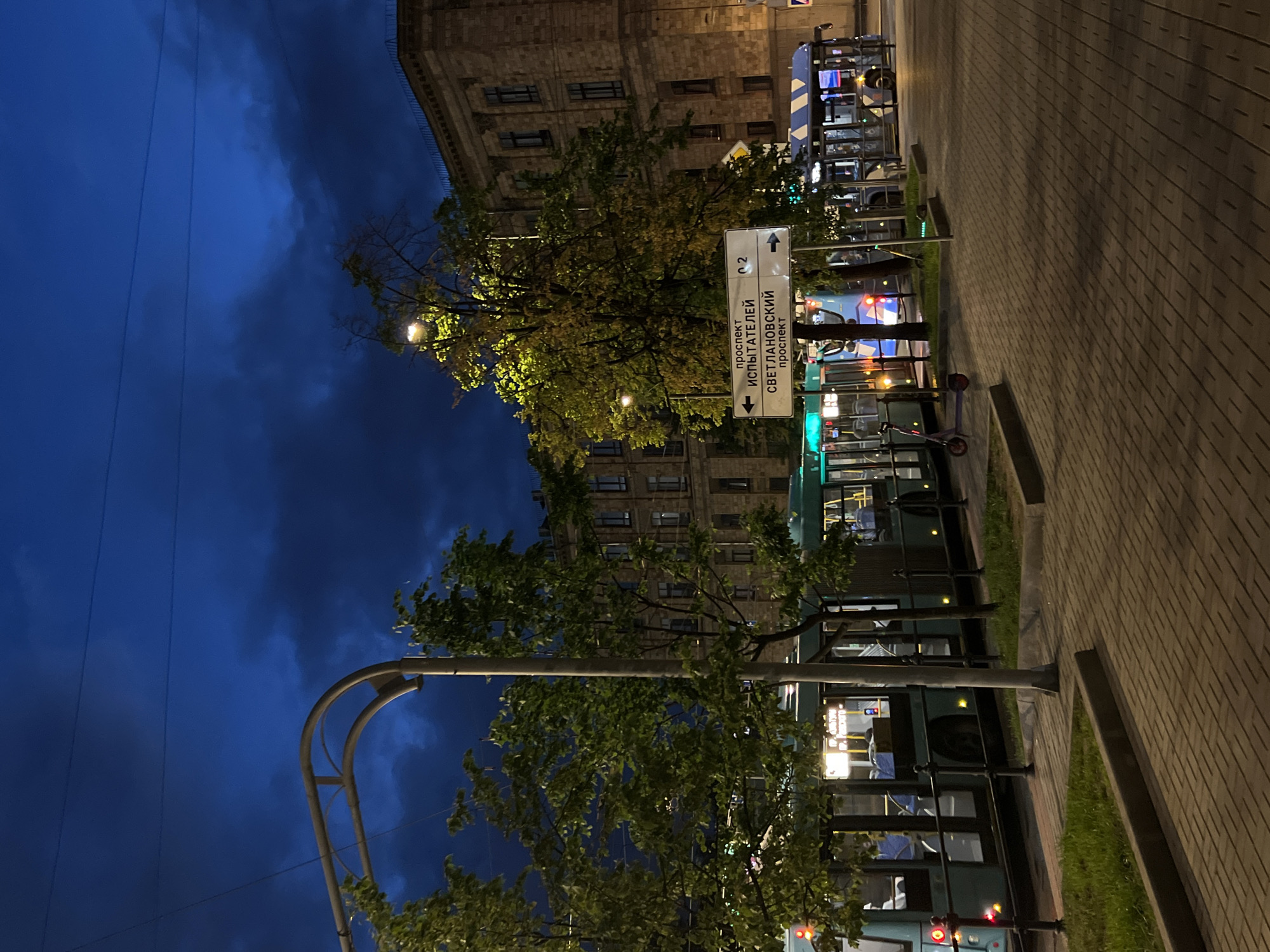 The image captures a nighttime scene on a city street in an unknown location in Russia. The sky is a deep, dark blue with dramatic clouds, suggesting twilight or a stormy evening. A street lamp illuminates a large tree with yellowing leaves, casting a warm glow.
In the foreground, a sidewalk paved with brick patterns leads towards a street where public transportation is present. Several buses are visible, one teal-colored and another with a blue and white striped design. One of the buses has its interior lights on, revealing passengers seated inside. The buses are parked along a curb, with black metal railings separating them from a grassy area.
A prominent white street signpost stands near the center of the image. The sign has Cyrillic text and arrows, indicating directions to "проспект Испытателей" (Prospekt Ispytateley) to the left and "Светлановский проспект" (Svetlanovskiy Prospekt) to the right. Two electric scooters are parked on the sidewalk near the signpost.
Behind the buses and trees, a building with stone facade and multiple windows is visible, its architecture suggesting an older, established urban area. The overall impression is one of a quiet, nocturnal urban setting.