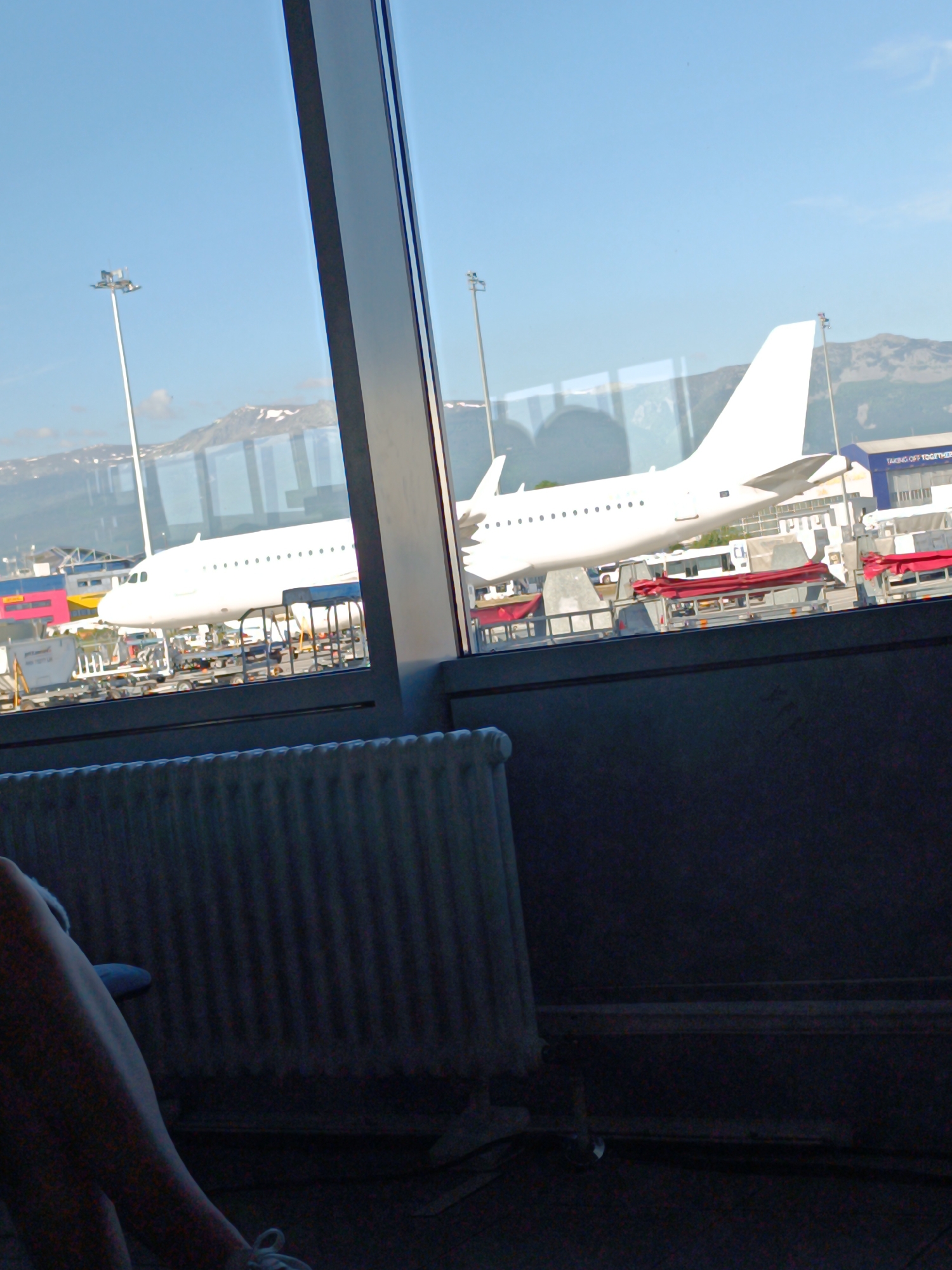The image, taken indoors, looks out of a window onto an airport scene. A white passenger airplane is visible on the tarmac, with various airport vehicles and equipment surrounding it. In the background, mountains can be seen. Inside, part of a radiator is visible in the lower left, with a person's leg and foot also present, suggesting the photograph was taken from an airport waiting area. Given the mountainous background, the likely location is Sofia Airport (SOF), Sofia, Bulgaria.