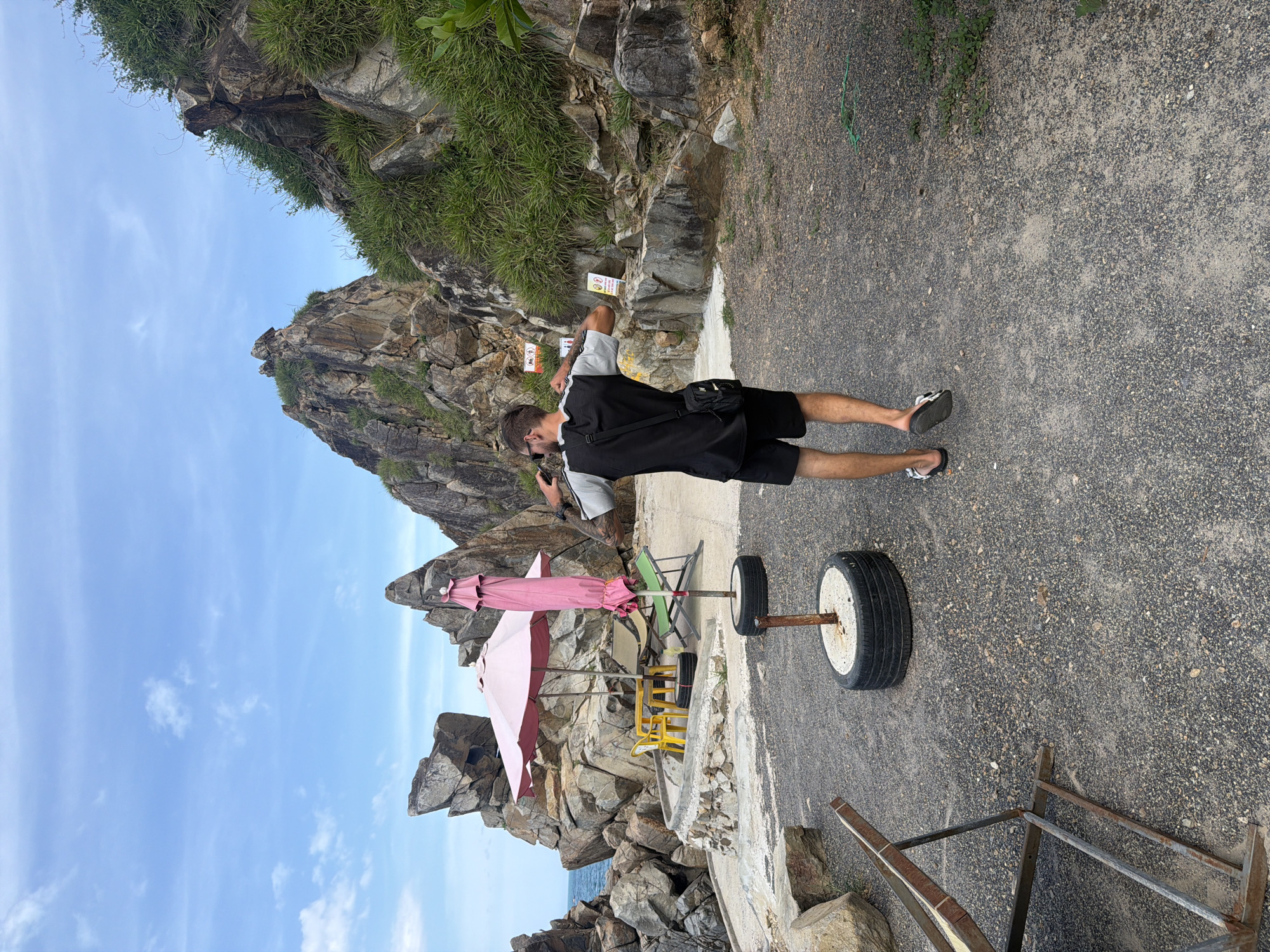 In Vĩnh Lương, Vietnam, a young man with sunglasses and a black and grey t-shirt, black shorts, and sandals is facing away from the camera, holding a phone up to his ear. He is standing on a gravel path in front of a rocky hillside. To his left, there are two pink umbrellas, one open and one closed, standing over a yellow table and chairs, and a green lounge chair. Behind these items is a large rock formation. In the foreground, there are two tires filled with concrete, serving as bases for poles. The sky above is blue with scattered white clouds.