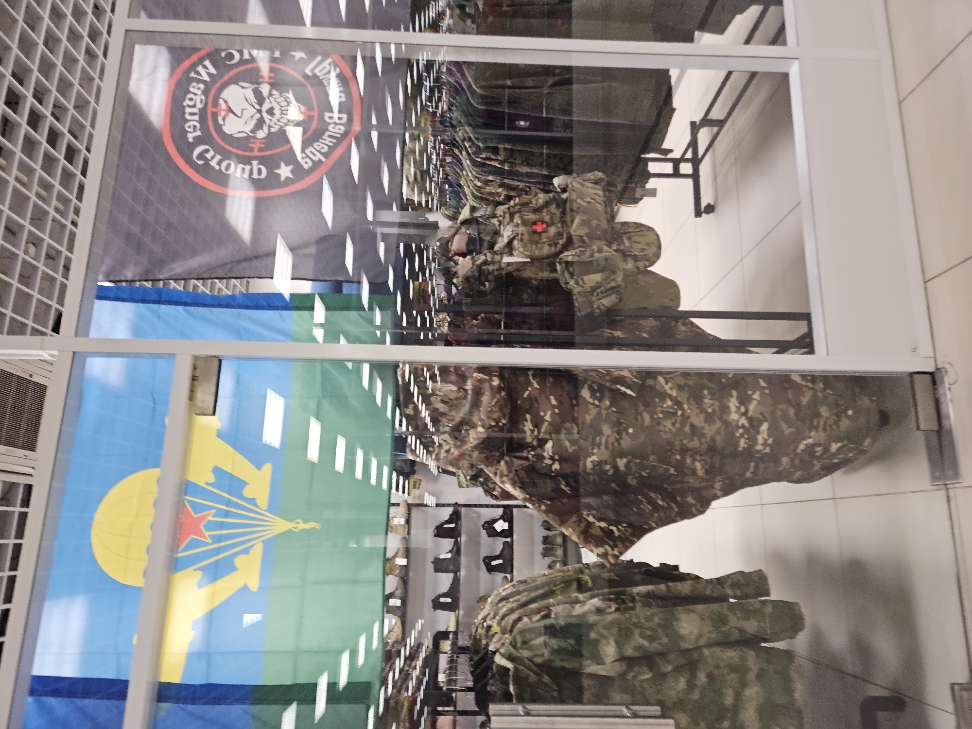 The image shows the storefront window of a military surplus or outdoor gear store in Ulyanovsk, Russia. Reflected in the glass are two large flags. The flag on the left is predominantly blue and green with a yellow parachute and a red star. The flag on the right is black with a circular emblem featuring a skull with crosshairs, and text in Cyrillic and what appears to be English. Inside the store, visible through the glass, are racks of camouflage clothing, display shelves with boots, and a mannequin or display stand dressed in tactical gear. The store appears well-lit with fluorescent lighting. The overall impression is of a retail environment selling specialized apparel and equipment.
