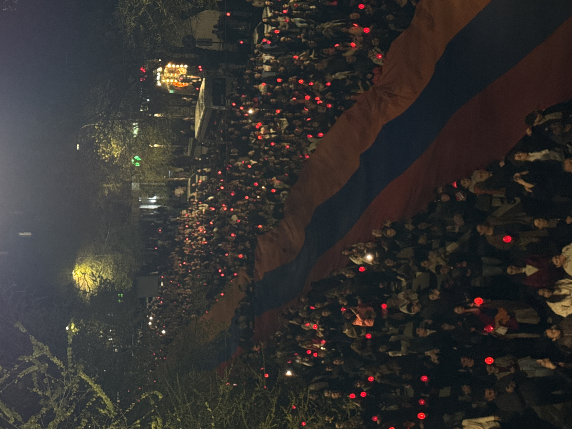 A large crowd of people is depicted from an elevated perspective, gathered outdoors at night in what is identified as Yerevan, Armenia. Thousands of individuals fill the frame, densely packed in a wide area.
The most prominent object is a very large Armenian flag, with its distinctive horizontal red, blue, and orange stripes, unfurled and extending across a significant portion of the crowd. Many attendees are holding small red lights, creating a scattered pattern of glowing red points throughout the assembly. These lights could be candles, phone flashlights with red filters, or small LED lights, adding a somber or unified atmosphere to the scene.
In the distance and along the left edge of the image, the soft glow of artificial lights from street fixtures and what appears to be a stage or an illuminated structure can be seen. Dark trees and sparse vegetation frame parts of the gathering. The event appears to be a large public demonstration, vigil, or commemoration, characterized by collective participation and a display of national identity. The time of day is clearly night. No visible text is discernible in the image.