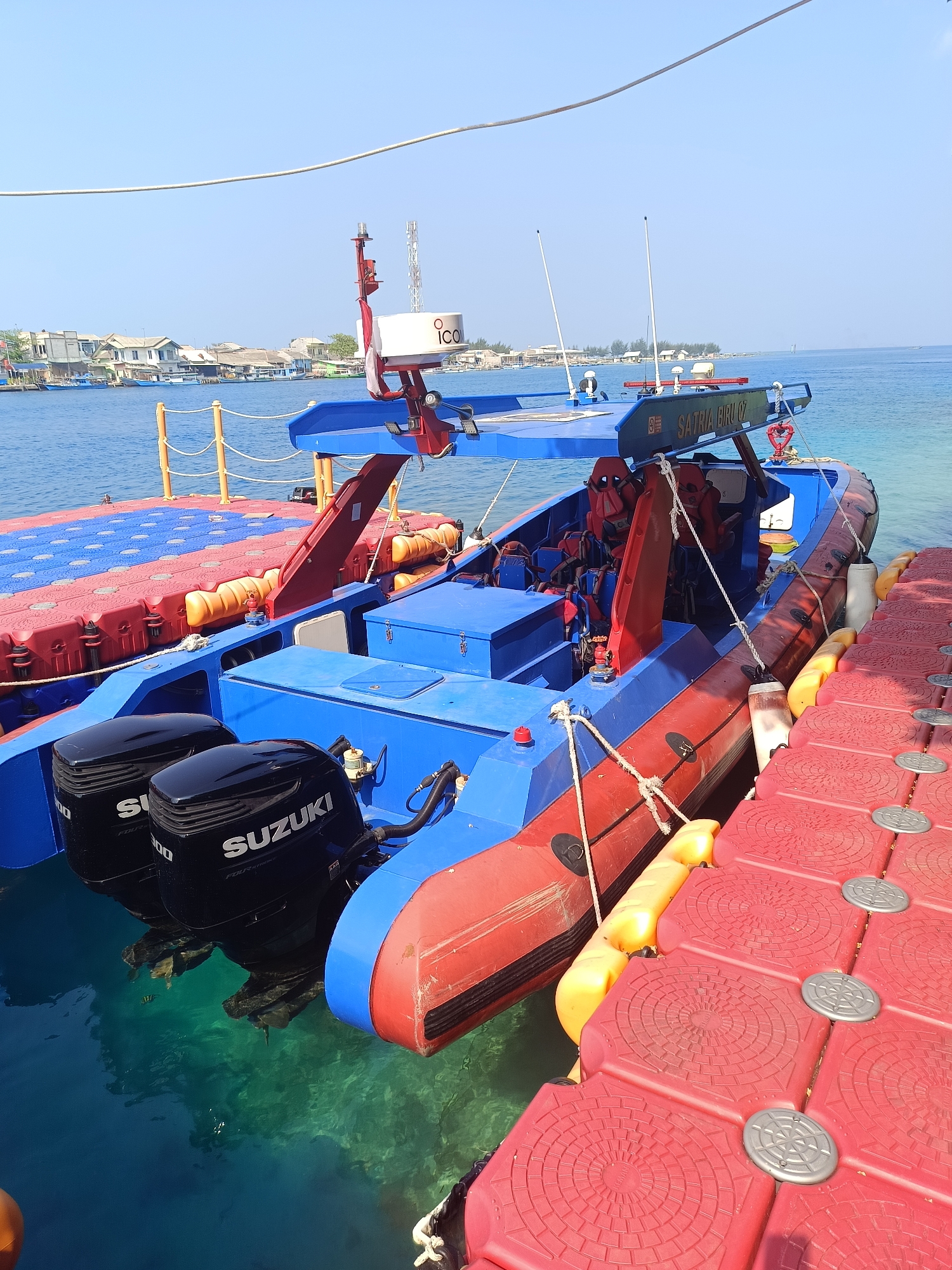 The image shows a blue and red rigid inflatable boat (RIB) docked at a red modular floating dock on a clear blue sea. The boat has two black Suzuki outboard engines mounted on the stern. Life vests are visible inside the boat, which is also equipped with a radar and antenna system. The boat is labeled "SATRIA BIRU 07". In the background, a coastline with houses and some vegetation is visible under a clear sky. The scene appears to be a sunny day. The location is the Special Capital Region of Jakarta, Indonesia, likely in one of its coastal areas or islands.