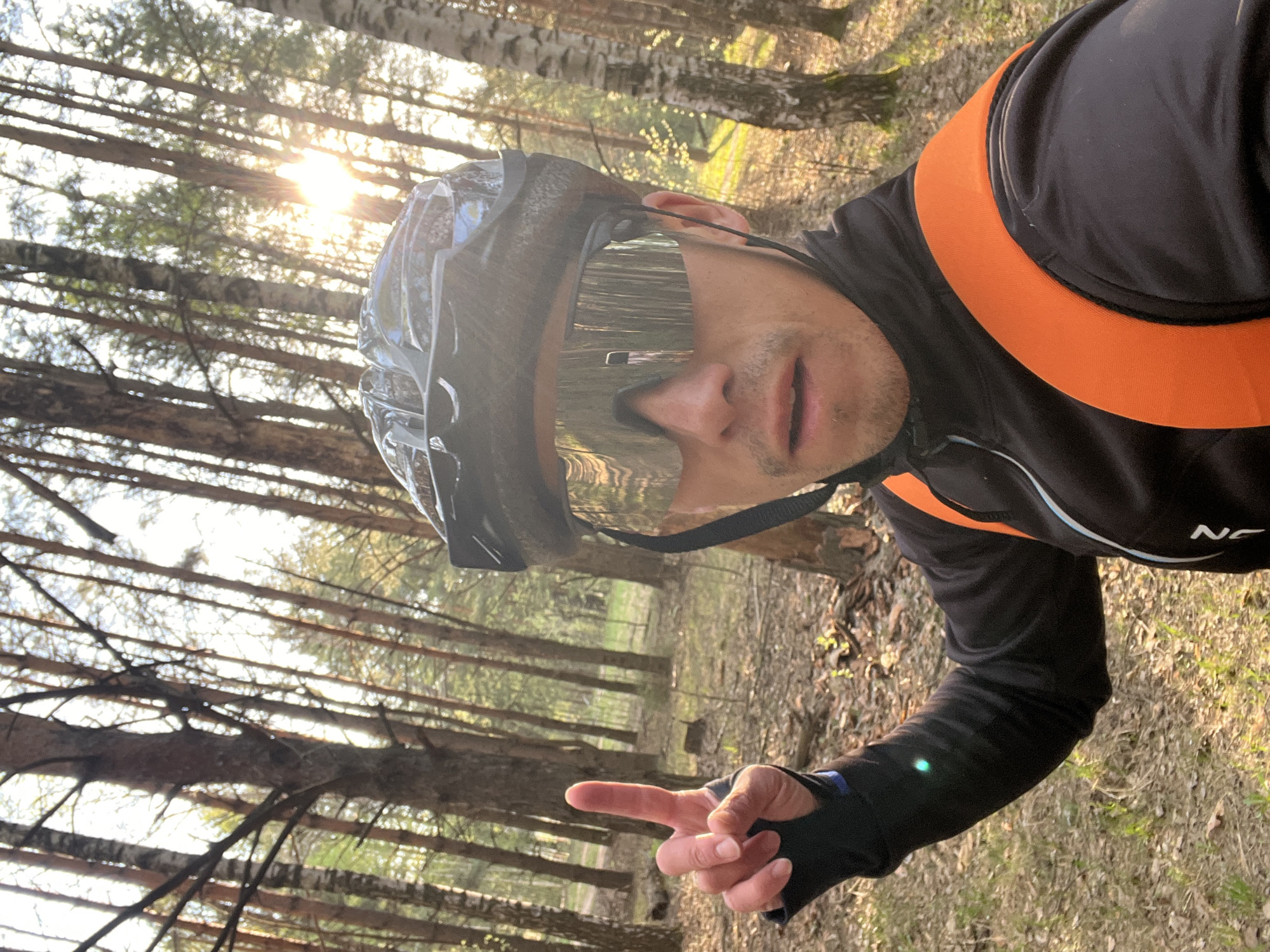 A selfie depicts a man wearing a black cycling helmet and mirrored sunglasses, positioned within a forest. He's dressed in a black, long-sleeved cycling jersey with an orange accent on the shoulders. The man is pointing towards the camera with his right hand. The background shows a dense forest with tall trees and sunlight filtering through the canopy. The location is Chaikovsky, Russia.