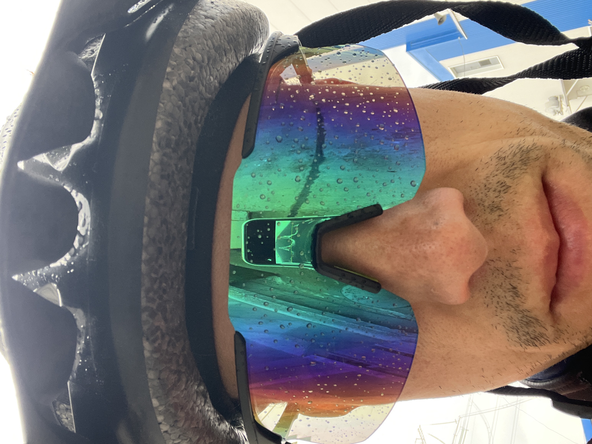 A close-up, low-angle shot captures a person's head, rotated ninety degrees to the right, giving the impression they are looking upwards. The individual is wearing a black cycling helmet with a textured surface and black straps. Prominently featured are large, wraparound sports sunglasses with iridescent or rainbow-colored lenses. These lenses are covered in numerous small water droplets, suggesting recent exposure to rain, sweat, or spray.
Visible parts of the person's face include their nose, mouth (slightly open), chin, and cheek, all covered with short stubble or facial hair. The person's expression appears neutral.
The background, visible above the person's head and through the sunglasses, indicates an indoor or sheltered setting. It shows a white ceiling with rectangular light fixtures or ventilation grates, some with a blue tint.
The presence of the cycling helmet and the water-splattered sunglasses strongly implies the person has recently engaged in an outdoor activity, likely cycling, during or immediately after wet conditions, possibly in Chaikovsky, Russia. There is no visible text in the image.