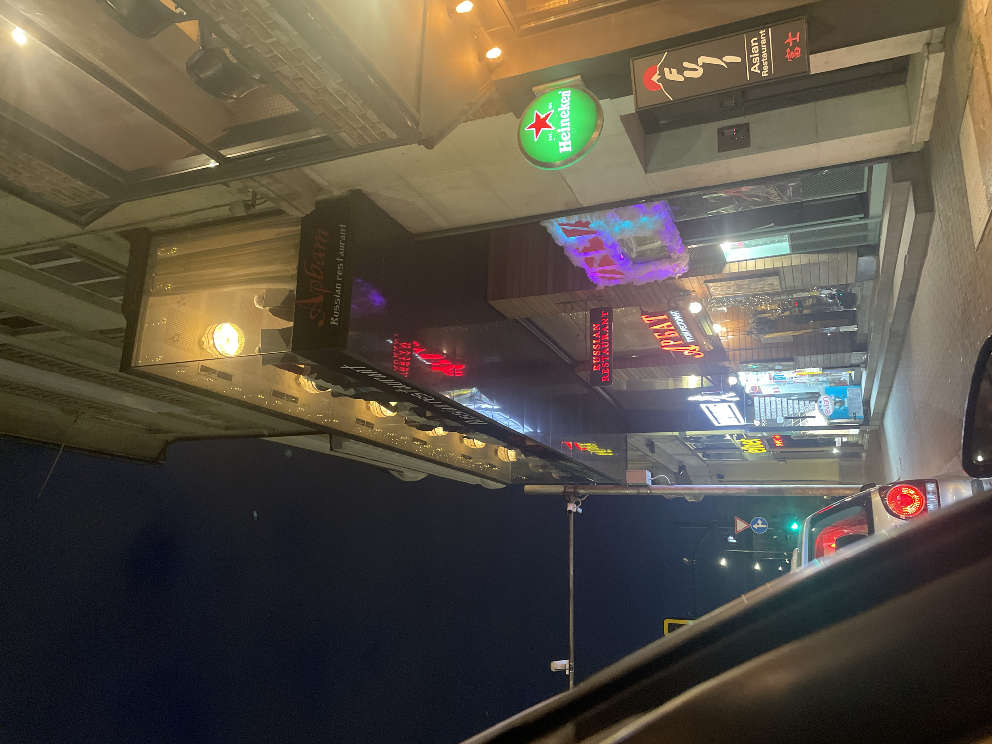 The image captures a nighttime street scene in Sofia, Bulgaria, viewed from inside a vehicle, with part of the car's window frame and dashboard visible in the foreground. The street is illuminated by various neon and lit signs, indicating a commercial area with multiple businesses.
On the left, a prominent dark sign displays "Арбат Russian restaurant" (Arbat Russian restaurant) in both English and Cyrillic script. Further along, another sign reads "RUSSIAN RESTAURANT" and "РУСКИ РЕСТОРАНТ" (Russian restaurant), suggesting another or the same Russian eatery. A bright green circular sign for "Heineken" with a red star is clearly visible. To the right, a sign for "FUJI Asian Restaurant 富士" identifies an Asian establishment. Below the Heineken sign, another illuminated display lists items such as "SOUVENIR, COFFEE, ESPRESSO, CAPPUCCINO, ICE CREAM, CHOCOLATE, COOKIES, WATER, PS, BEER, NUTS, TEA, evian," indicating a small shop or kiosk.
The sky is dark, characteristic of nighttime. A red taillight of a vehicle and a green traffic light are visible in the distance, suggesting traffic movement or a stop. Reflections from the illuminated signs and streetlights are cast onto the wet or glossy surfaces of the buildings and possibly the car's window. The overall scene depicts a bustling urban environment after dark, characterized by diverse culinary offerings and commercial activity.