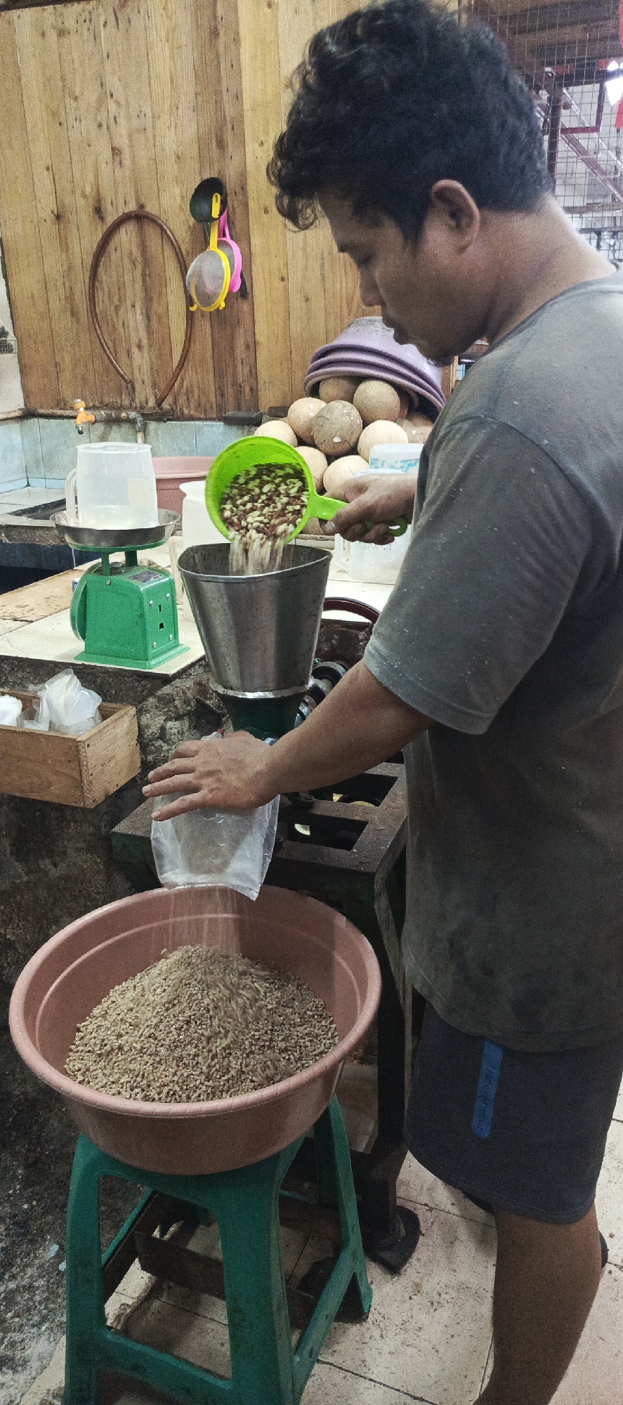 A person is standing in what appears to be an outdoor market stall in Sukaresmi, Indonesia, working with a grain processing machine. The person, wearing a dark grey t-shirt and dark shorts, is actively pouring a scoop of mixed grains into a funnel attached to a green and silver machine. Below the machine, a large, pink plastic bowl is positioned on a green plastic stool, and a stream of processed grain is being poured into it from a clear plastic bag held by the person. In the background, a wooden wall with various items hanging on it, including a brown hoop and a colorful cleaning tool, suggests a rustic setting. Other objects visible include a green scale with a measuring pitcher, a wooden box containing smaller bags, and a pile of large, round items that could be coconuts or gourds. The floor is tiled and appears to be slightly wet in some areas. The overall scene depicts a common marketplace activity involving food preparation or processing.