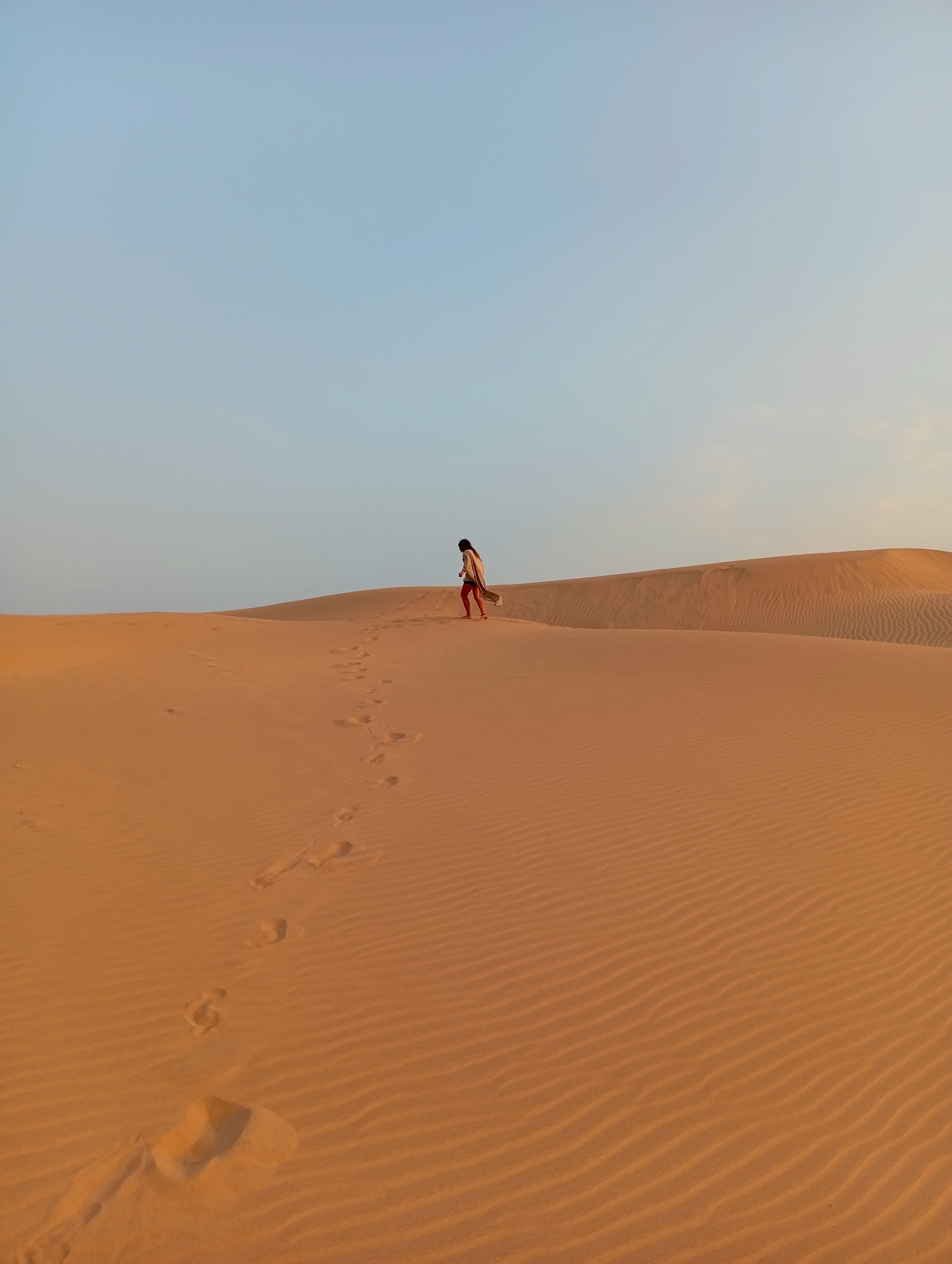 A full shot captures a person walking across a large sand dune in Phuoc Dinh, Vietnam. The person is small in the frame, positioned on the upper portion of the dune, with footprints leading away from them down the slope. The sand is rippled with wind patterns. The sky is clear and light blue.