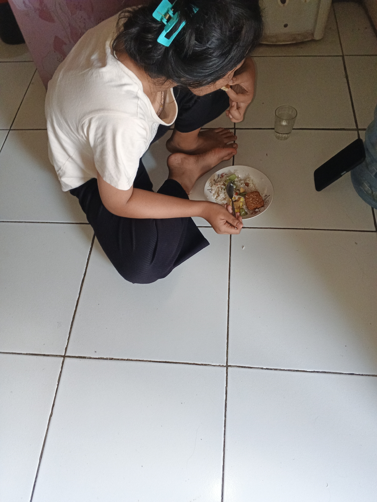 An overhead shot shows a person sitting on a tiled floor, eating a meal. The person is wearing a white t-shirt and dark pants, and their hair is pulled back with a turquoise clip. They are holding a spoon in their right hand, bringing food to their mouth. A plate of rice with side dishes is in front of them, along with a glass of water and a smartphone. The floor is white tile with dark grout lines. The background includes part of a pink patterned object and a white appliance. The scene appears to be indoors, likely in a residential setting in Warungkiara, Indonesia.