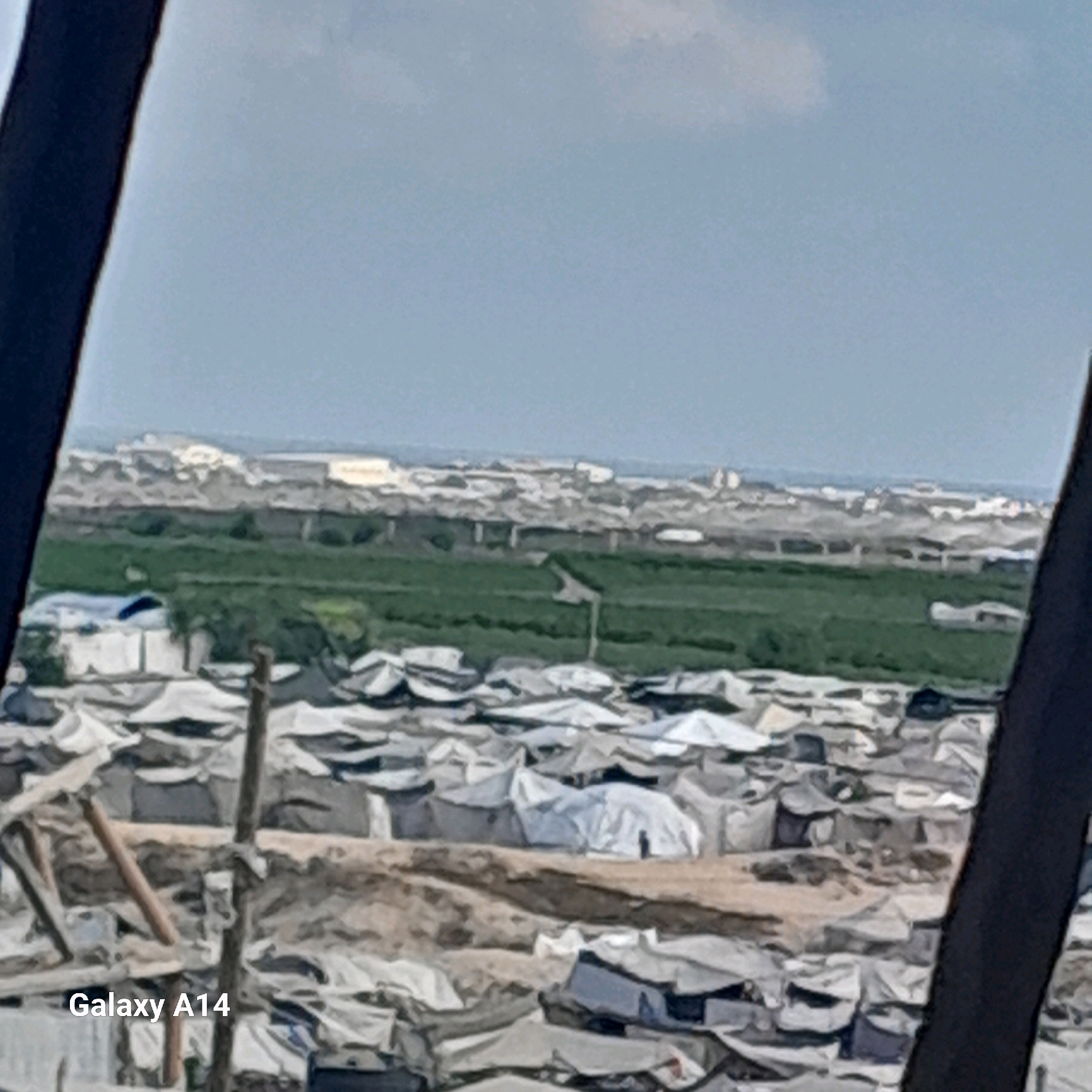 The image shows a densely populated camp with numerous tents and temporary shelters. In the foreground, a wooden pole stands vertically, with a dark, thick vertical element on the right edge of the frame. The tents are predominantly white and gray, with some showing signs of wear and tear. The ground appears to be dusty and uneven. In the midground, there is a strip of green fields, possibly agricultural land, with some structures visible behind it. The background depicts a more urban landscape with buildings and what appear to be larger structures or industrial facilities. The sky is overcast with a light blue hue. The word "Galaxy A14" is watermarked in the bottom left corner of the image. This scene is consistent with the visual appearance of refugee camps in Khan Yunis, Palestinian Territories, which have experienced significant population displacement and the establishment of temporary settlements.