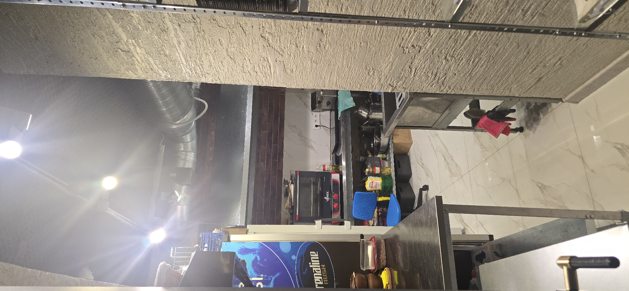 The image shows a commercial kitchen area in Krasnodar, Russia. The scene is captured from a slightly elevated perspective, looking into the kitchen from what appears to be a doorway or aisle.
The kitchen is equipped with stainless steel countertops and shelving. On a shelf, there is a black oven with a red trim, labeled "VENIX". To the left of the oven, there's a large blue plastic crate. Below the shelves, black storage bins are visible.
In the foreground, a stainless steel table occupies the left side of the frame, with a brushed metal handle visible on its edge. To the right, another stainless steel work area is present, featuring a sink basin. A red cloth and a brush are draped over the leg of this work area.
The walls are textured, with one side appearing to be a rough, light-colored stucco and the other a dark brick pattern. The floor is tiled with a light-colored marble pattern. Overhead, metallic ductwork is prominent, along with bright lighting fixtures that create glare.
On one of the walls, there is a large blue poster with text that includes "renaline RUSH". Various kitchen supplies and utensils are scattered throughout the scene, including pots, pans, bottles, and containers. There is no visible activity or people actively engaged in cooking or working in the kitchen. The overall impression is that of a functional, albeit somewhat cluttered, commercial kitchen environment.