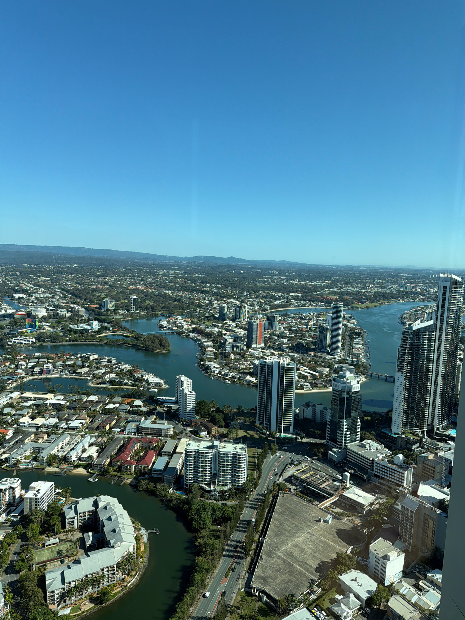 This is an aerial, high-angle, wide shot of a coastal city on a clear, sunny day. The expansive blue sky dominates the upper half of the image. Below, the city is a dense tapestry of buildings, waterways, and greenery, extending to a distant hazy mountain range. The architecture is varied, featuring numerous high-rise apartment buildings with reflective glass facades, as well as lower-density residential areas with red-tiled roofs and courtyards. Several canals and a wider river system weave through the urban landscape, dotted with boats and jetties. A prominent bridge spans one of the waterways. The foreground shows a particularly dense cluster of modern high-rise buildings and a complex of low-rise buildings with a curved design, surrounded by water and trees. Further back, the cityscape transitions into more suburban sprawl with houses and trees, eventually merging with the natural landscape of hills and mountains in the distance. The overall impression is of a developed coastal urban environment with significant water features and clear weather. The location is the city of Gold Coast, Australia, with the distinctive skyline and canal system indicative of the Broadbeach or Surfers Paradise area.