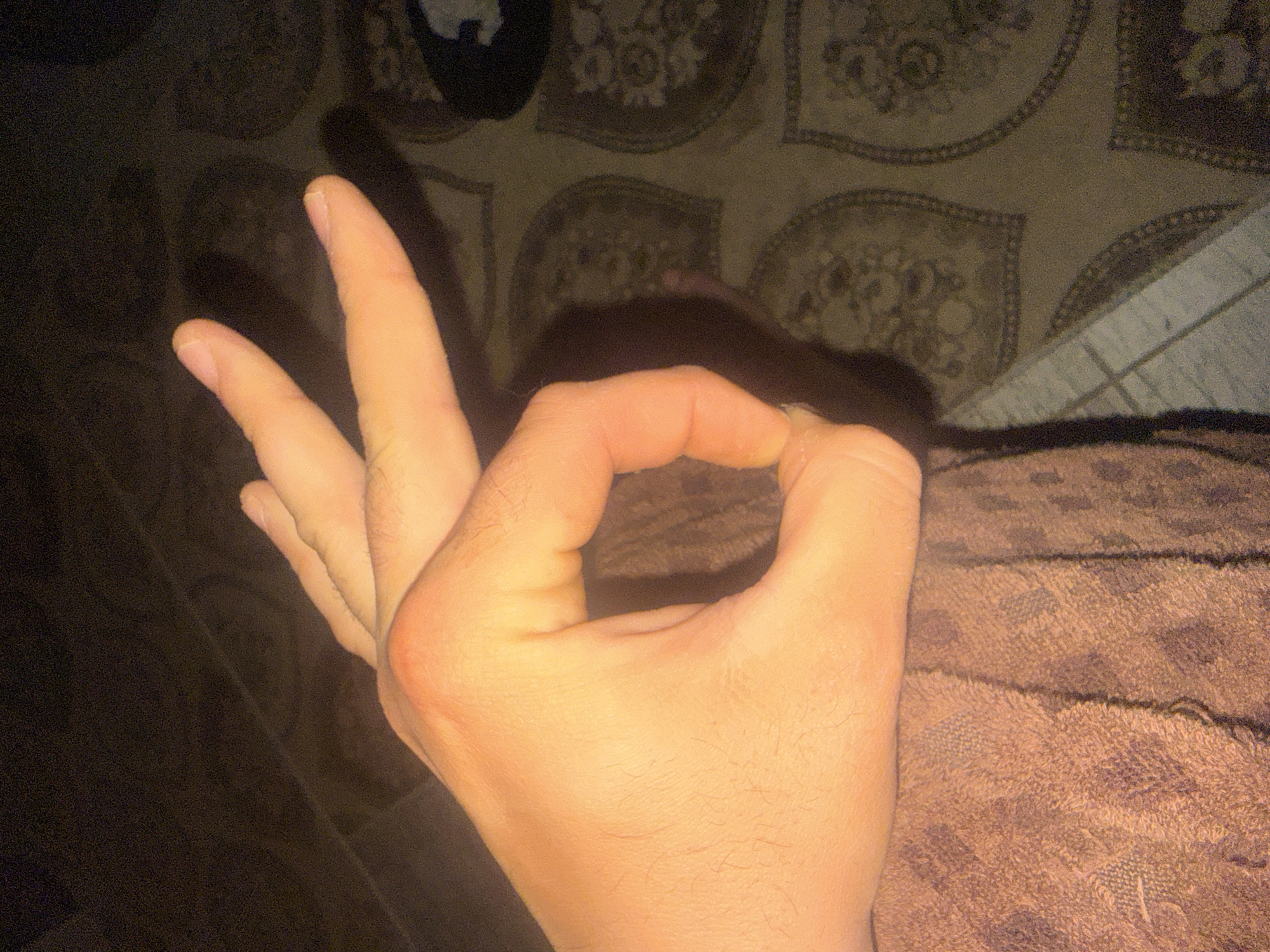 A human hand is making an "OK" gesture against a background consisting of bedding with various patterns. The thumb and index finger form a circle, while the other fingers are extended upwards. The hand's skin is fair in tone. The surrounding environment is dimly lit, with varying textures and designs on the bedding.