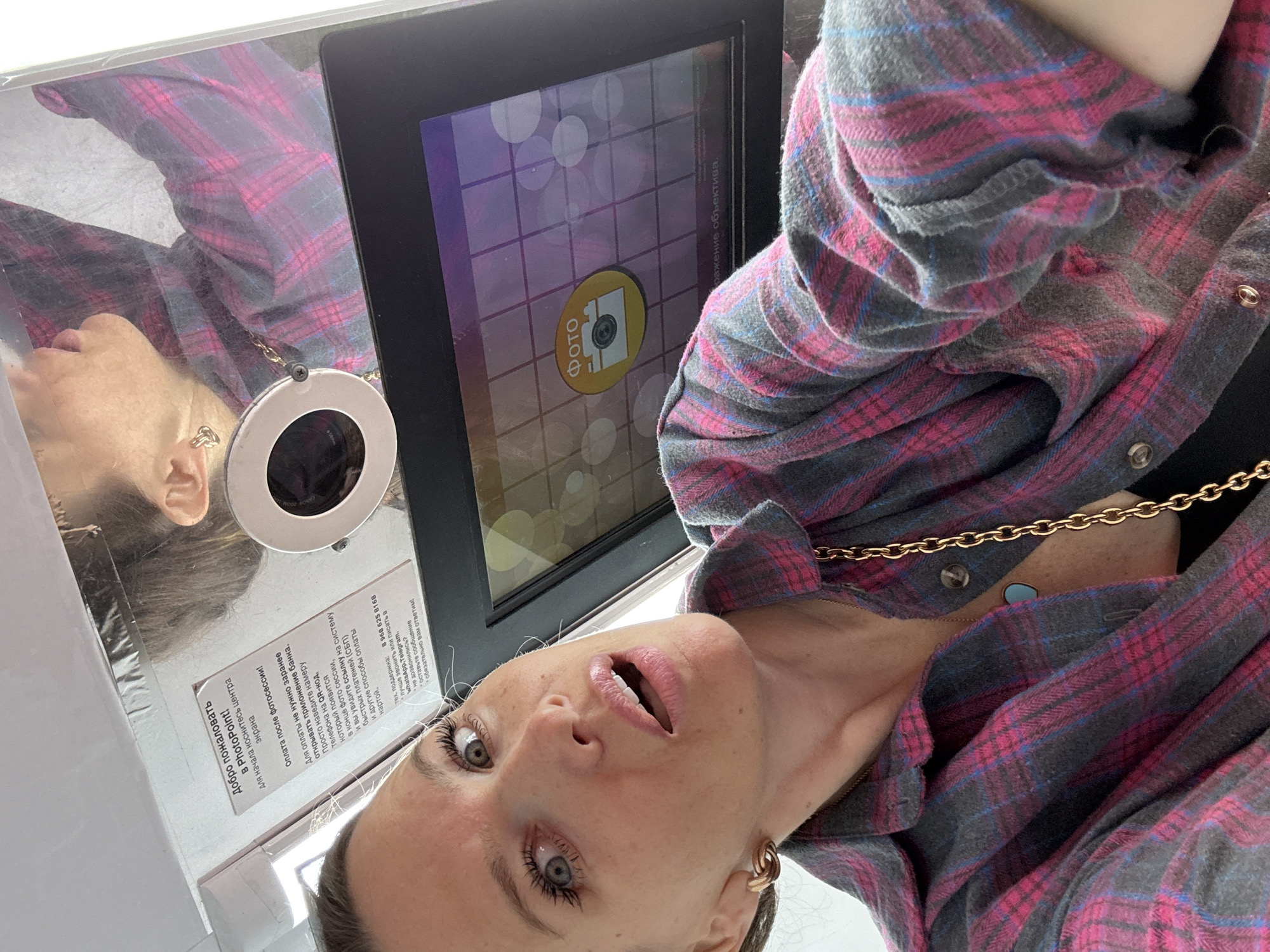 A woman with blonde hair and light skin takes a selfie in a photo booth. She is wearing a pink and gray plaid shirt over a black top, and a gold chain necklace with a turquoise pendant. She is looking at the camera with her mouth open. The photo booth has a screen with a "Photo" button and a circular lens with a light ring. A reflection of the woman and the photo booth can be seen on a mirrored surface above her. A sign in Russian provides instructions for using the photo booth. The background is blurred, but appears to be indoors. The scene is likely within Moscow, Russia, given the language on the sign.