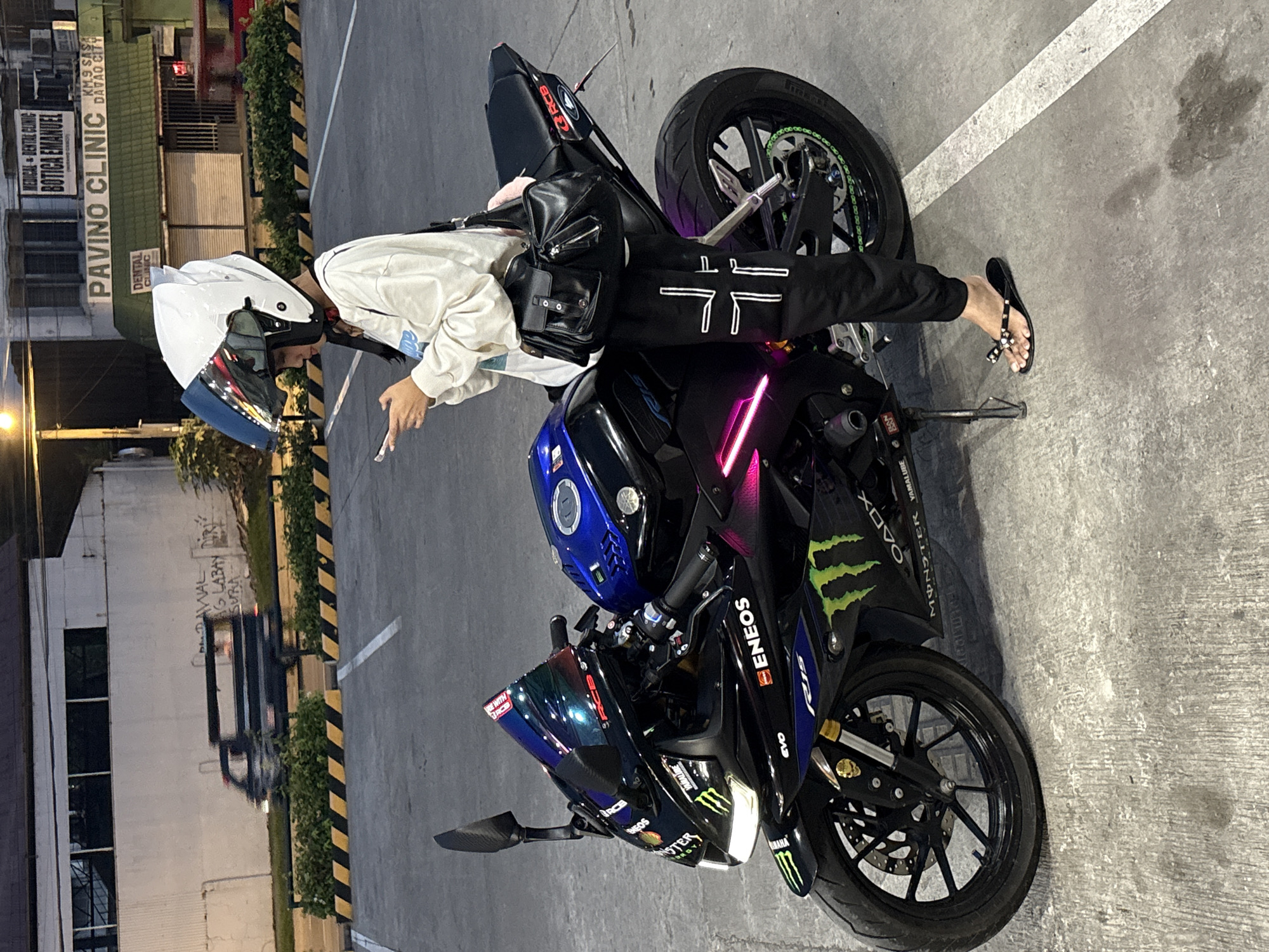 In Davao City, Philippines, a young person wearing a white helmet with a blue visor and a white long-sleeved shirt is standing next to a black Yamaha R15 motorcycle. The motorcycle is adorned with numerous decals, including prominent "Monster Energy" logos and "ENEOS" branding. The person is looking at their mobile phone, held in their left hand, while their right hand rests on the motorcycle's seat. They are wearing black pants with a white cross design and black flip-flops. A black shoulder bag with multiple compartments is slung over their right shoulder. The motorcycle is parked on a concrete surface, marked with yellow and black diagonal lines, characteristic of parking lot delineations. In the background, a building with signage for "PAVINO CLINIC" and "MEDICAL - DENTAL CLINIC BOTICA EMANUEL" is visible. The scene appears to be an outdoor parking area, possibly at dusk, given the lighting conditions.