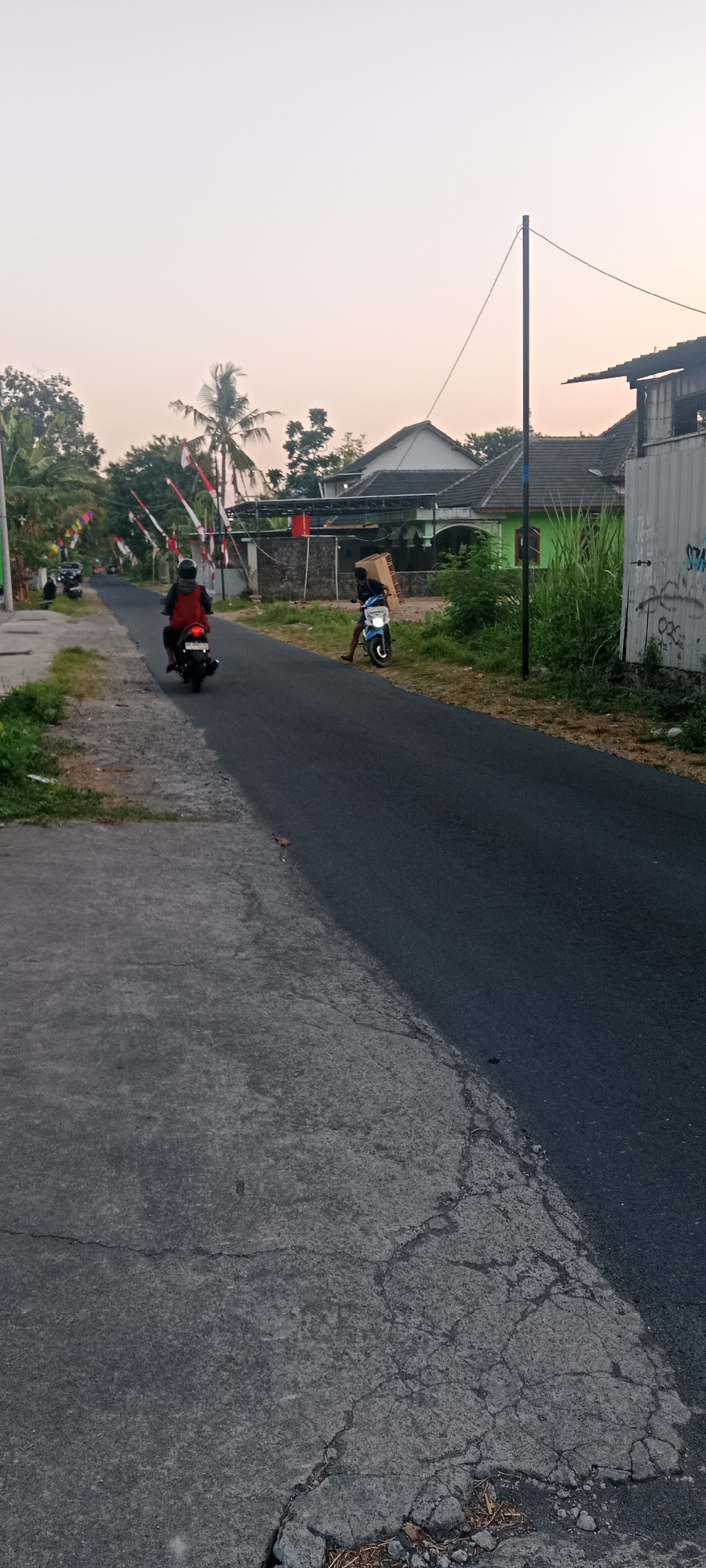 In Maguwoharjo, Indonesia, this image captures a street scene during what appears to be late afternoon or early evening. A paved road runs through a suburban or rural area, flanked by a concrete sidewalk on the left and vegetation on the right. Several Indonesian flags, red and white, are strung along poles on the left side of the road, indicating a local celebration or national holiday.
On the road, two individuals are depicted on motorcycles. Closer to the foreground, a person wearing a red vest and helmet is riding a black motorcycle away from the camera. Further down the road, a second person on a blue motorcycle is stationary, with a large cardboard box balanced on their shoulder, appearing to be moving the box. The environment includes scattered houses with visible roofs and walls, some with fences and lush green foliage. A tall, dark pole with a wire extending from it stands on the right side of the road. The sky is a pale, hazy orange, characteristic of dusk. The road surface shows signs of wear, with cracks and patches visible on the concrete sidewalk.