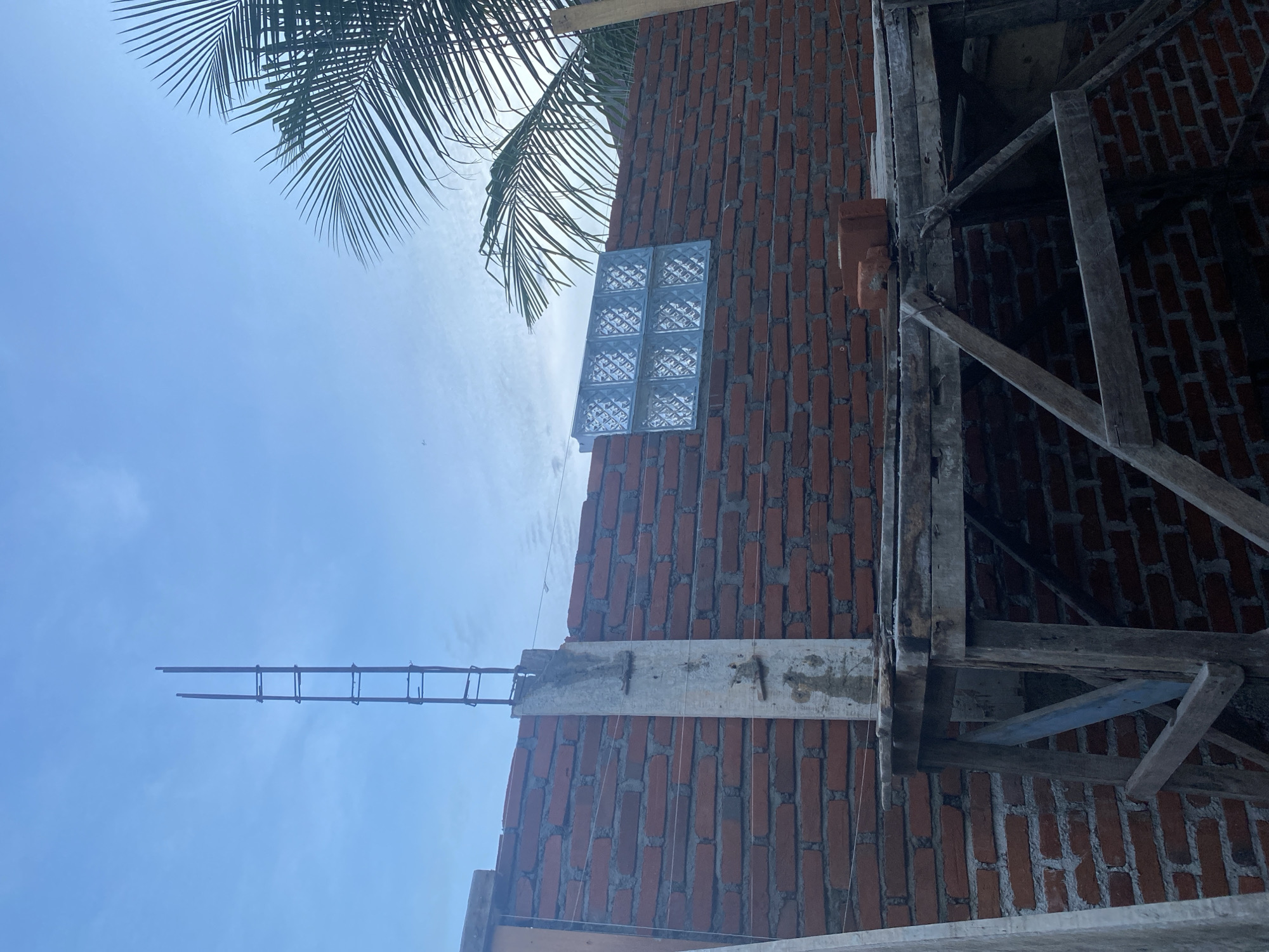 This image depicts an outdoor construction scene. A brick wall, partially constructed, dominates the frame, with a concrete column and exposed rebar rising vertically from its surface. A set of glass blocks is installed within the brickwork, forming a small window. Wooden scaffolding is positioned in front of the wall, supporting planks and construction materials. A palm frond extends into the upper right corner of the image, suggesting a tropical or subtropical environment. The sky is overcast with a light blue hue. The scene is devoid of any individuals. The location of Banda Aceh, Indonesia, is implied by the context.