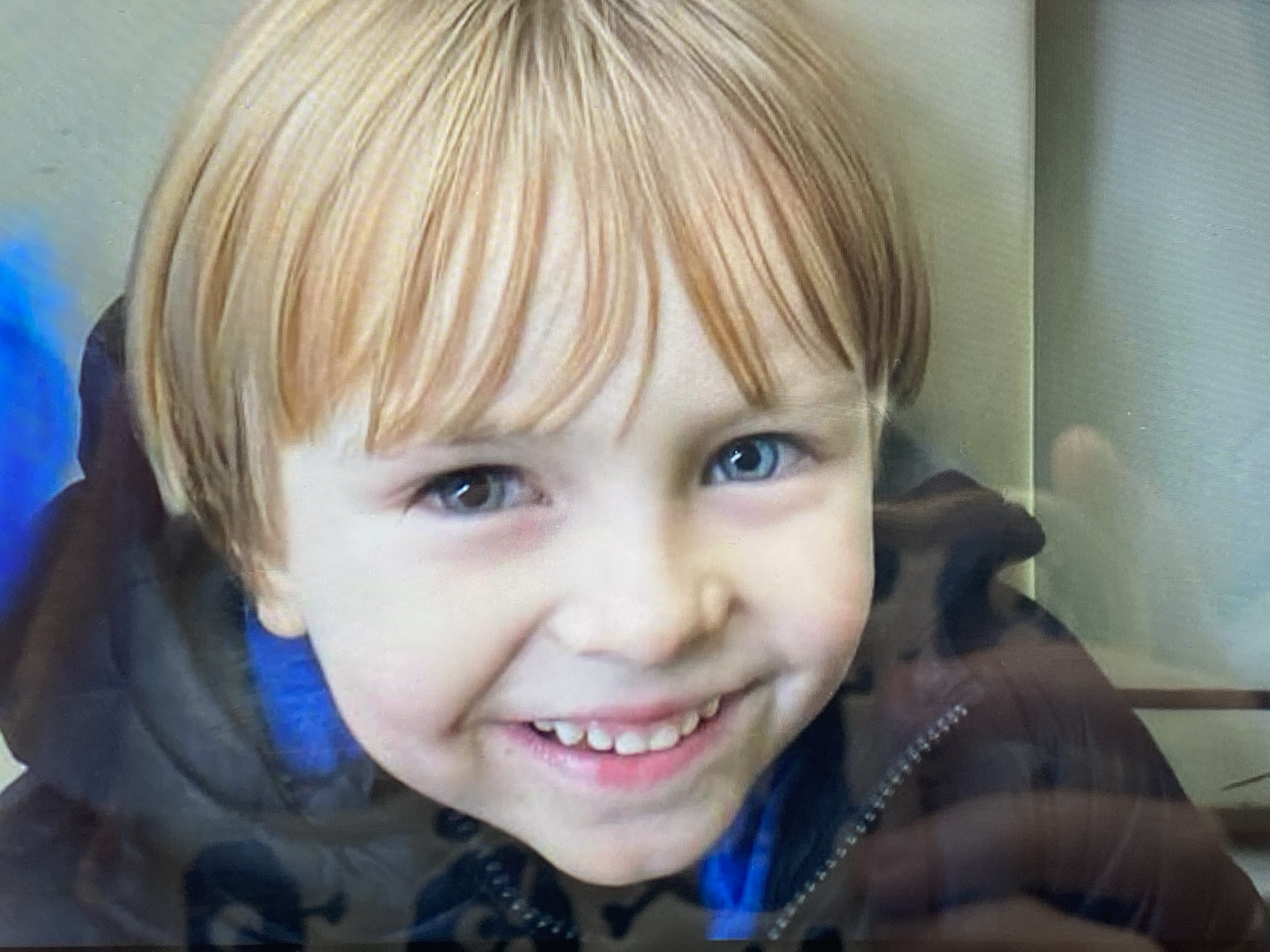 A close-up, head-and-shoulders photograph captures a young boy with blonde hair and a blue jacket. His hair is neatly parted and falls across his forehead. He has heterochromia, with one eye appearing blue and the other a darker shade, possibly brown or hazel. A broad smile reveals his teeth. He is wearing a dark hooded jacket over a bright blue layer underneath. The background is indistinct, appearing as a plain, light-colored wall. The image was taken in Pivdennoukrainsk, Ukraine.