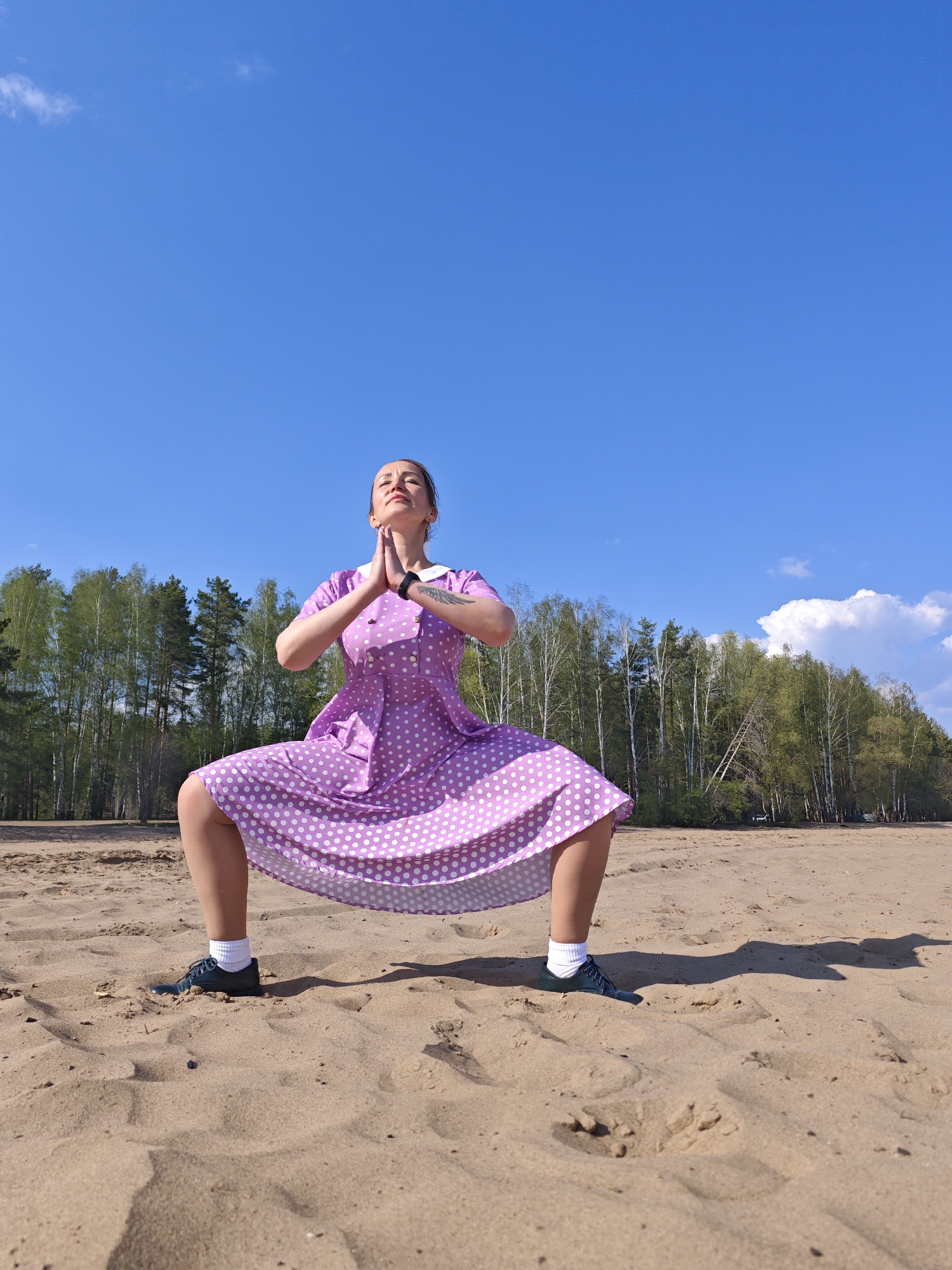 A woman is performing a yoga pose on a sandy beach. She is wearing a light purple polka dot dress, white socks, and dark shoes. Her hands are pressed together in front of her chest, and her eyes are closed. The sky is blue with a few scattered clouds, and a line of green trees can be seen in the background. The location is Noviy, Russia.