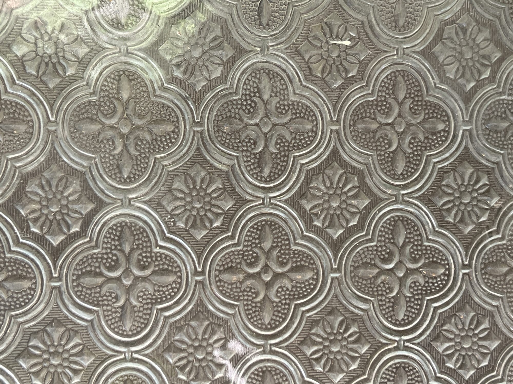 A close-up, overhead shot reveals a textured, patterned glass surface. The glass features a repeating ornate design that gives it a decorative, translucent appearance, diffusing any light passing through.
The primary motif is a series of interconnected quatrefoil shapes, each with four lobes. Within the center of each quatrefoil, there is a symmetrical, scroll-like or floral design. The lobes of these quatrefoils are filled with a fine, textured pattern, resembling small bumps or stippling. These quatrefoils are bordered by raised, smooth lines that define their shape.
Between the main quatrefoil patterns, there are smaller, roughly square sections. These sections typically contain a distinct floral design, appearing as a multi-petaled flower with a central point, surrounded by simpler geometric textures like diagonal lines. The entire pattern is embossed or etched into the glass, creating a three-dimensional effect.
The material itself appears to be clear glass, but the intricate patterning and texture scatter light, creating a greyish-white tone with varying shades that highlight the raised elements of the design. The background visible through the glass is very blurred and indistinct, suggesting a distant or out-of-focus scene, possibly with some green hues indicating foliage.
There are no people, objects other than the glass, or any visible text in the image. No specific activity is taking place; it is a static detail shot of a decorative surface, possibly part of a window, door, or partition.