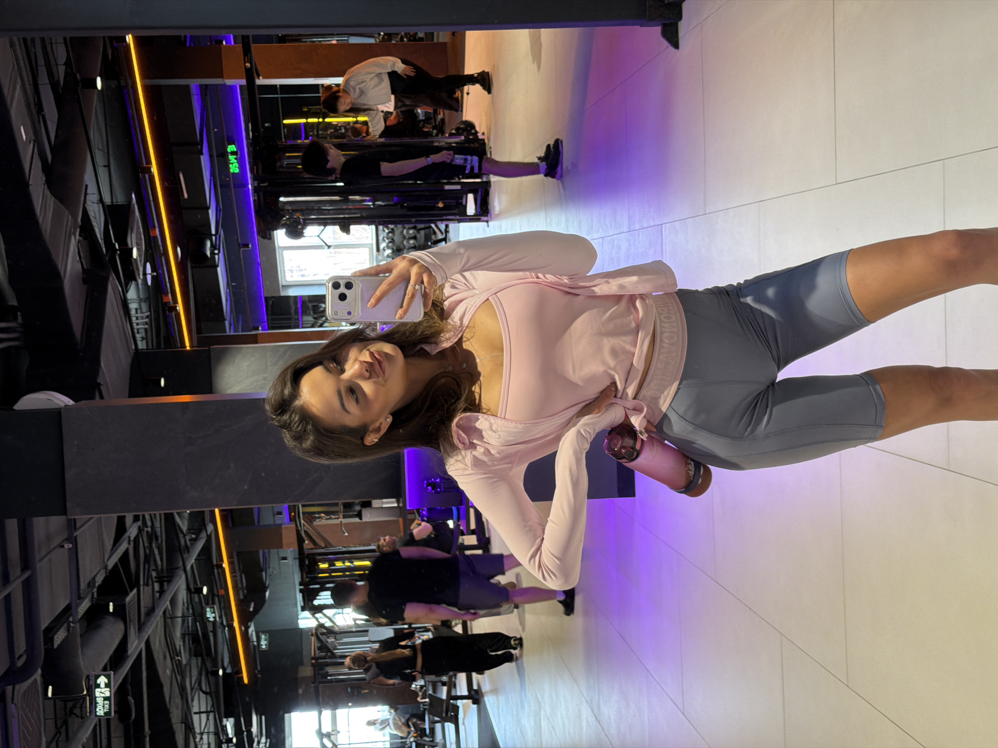 The image captures a young woman in what appears to be a fitness center, likely in Krasnoyarsk, Russia, based on visual cues. She is positioned facing the camera, holding a smartphone, presumably taking a selfie. She is wearing athletic attire consisting of grey cycling shorts and a pink long-sleeved top. A water bottle is held in her other hand.
The background shows an operating fitness center with gym equipment. There are other individuals working out in the background. The gym has modern design elements including colored lighting, and mirrors.
