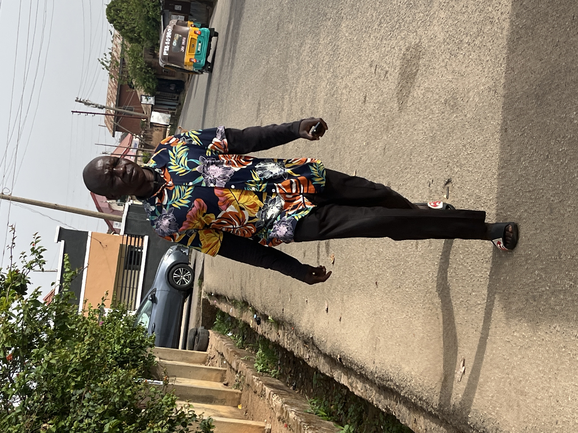 A man stands on a paved street in Jos, Nigeria. He is wearing a floral print short-sleeved shirt over a long-sleeved black undershirt, black pants, and sandals. The man appears to be holding a cell phone in his right hand.
Behind him is a parked gray sedan. Beyond the sedan is a building with a tan-colored exterior and a black section. To the left, there is a bush. A "Keke Napep" three-wheeled vehicle is parked at the end of the road. Power lines are visible overhead. The weather appears sunny.