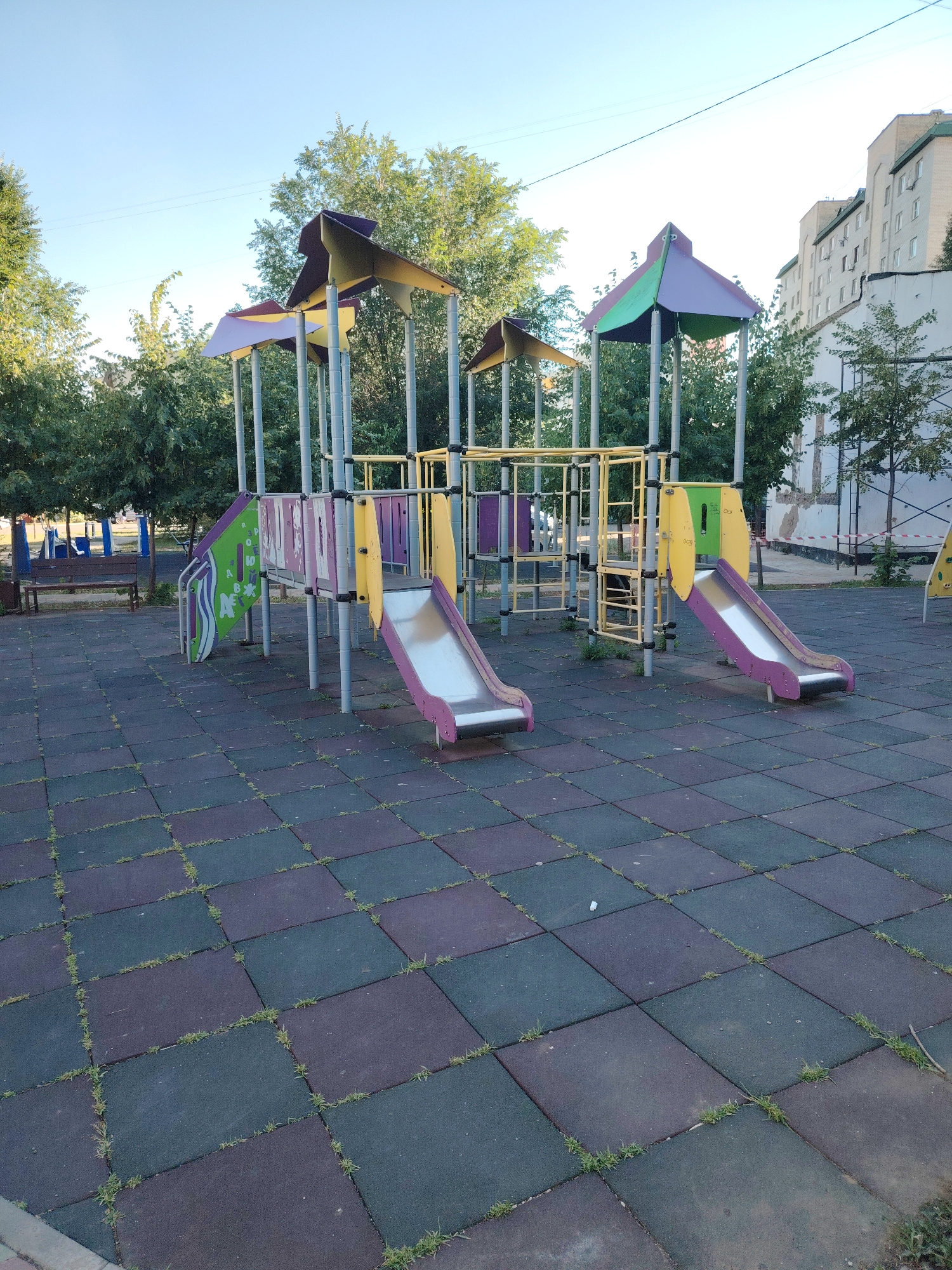 The image depicts an outdoor children's playground in Astana, Kazakhstan. The playground features a multi-level play structure with two purple and green roofed sections, supported by gray metal poles. Attached to the structure are two slides: one is purple with yellow railings and leads to the ground, and another is green with purple elements and a gray slide. The ground surface is made of interlocking rubber tiles in shades of dark blue, purple, and green, with grass growing between some of the tiles. In the background, there are trees and a multi-story residential building with a light-colored facade and green trim. A wooden bench and a trash can are visible to the left of the playground structure, along with other playground equipment in the distance. The sky is clear and blue.
