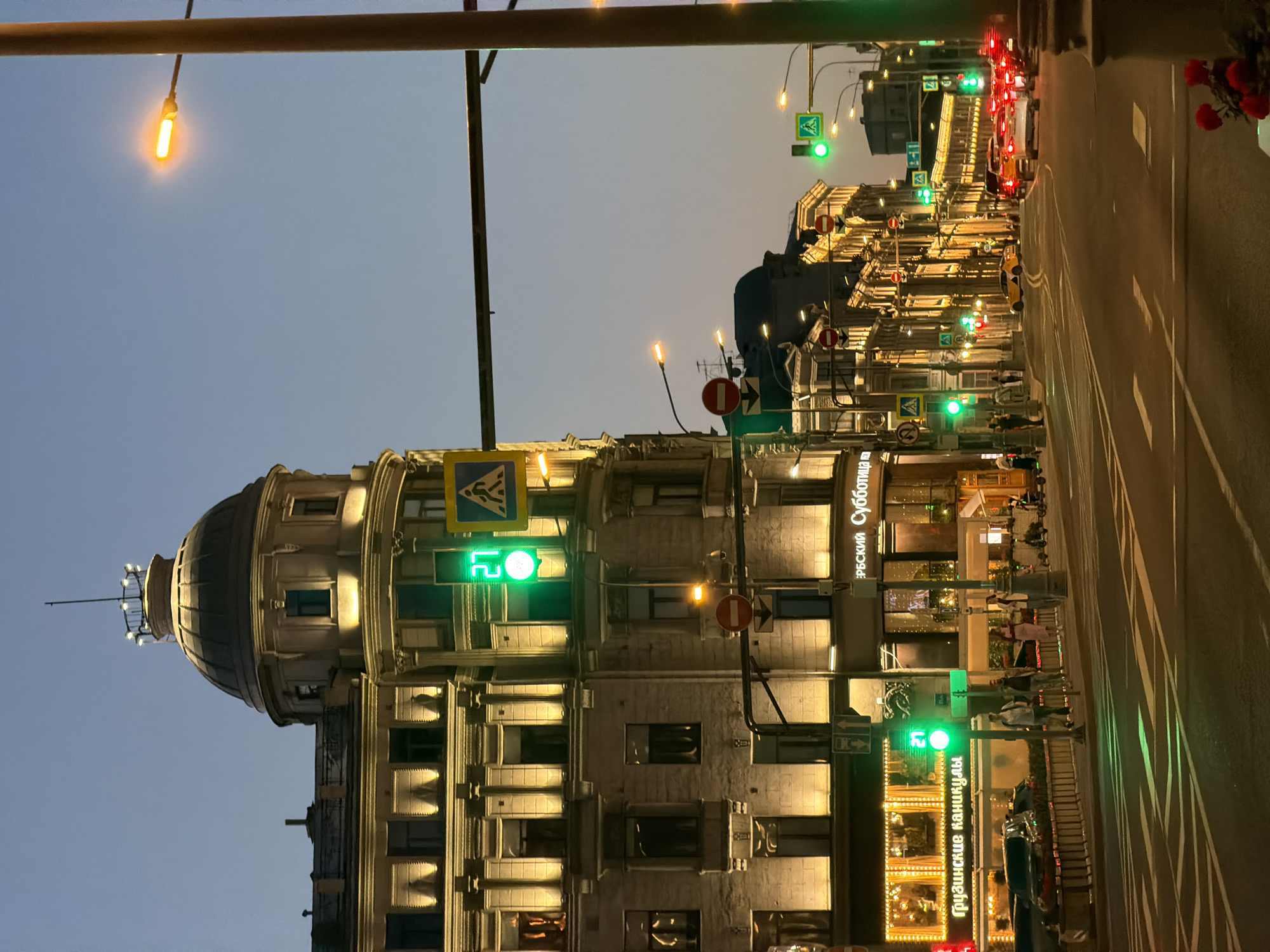 This photograph captures an urban scene in Moscow, Russia at dusk or early evening. The image is oriented sideways, with the main focus being on a multi-story building featuring a prominent dome on its corner. The building is illuminated with warm lighting, accentuating its architectural details.
A street runs along the right side of the frame. The street is marked with lane lines and lined with buildings featuring illuminated storefronts and signs written in Cyrillic. There are traffic lights visible, most of which are green.
Traffic is present on the street, including cars, and pedestrians are crossing the street. Above the buildings, the sky transitions from a pale blue near the horizon to a darker blue higher up. A pedestrian crossing sign is prominently displayed along with no entry signs.