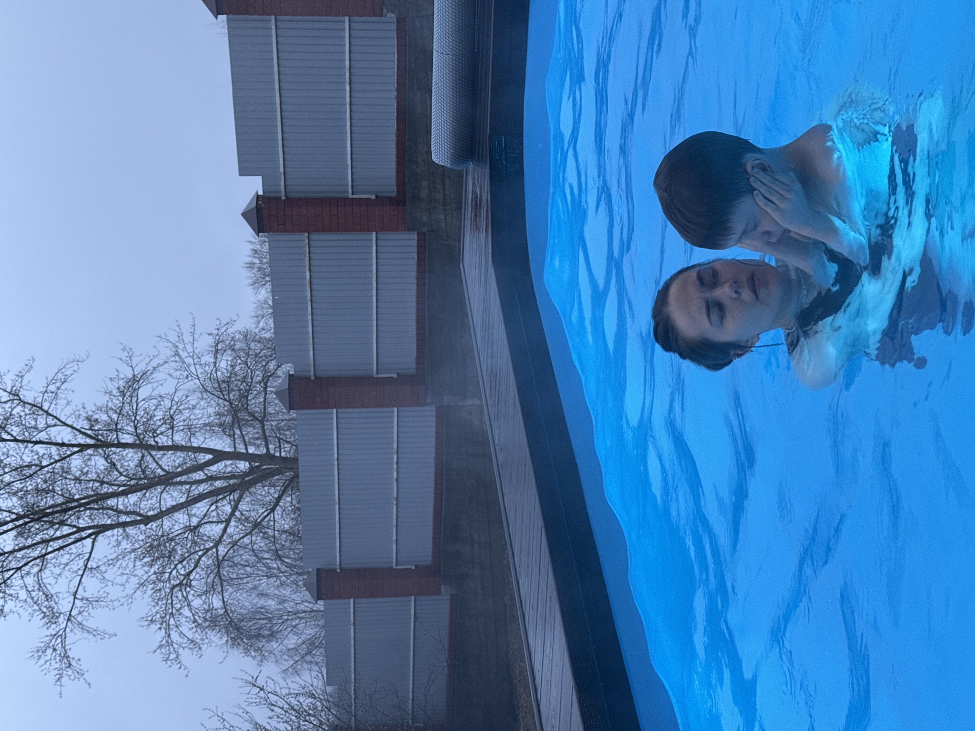 A woman and a child are in a swimming pool. The pool is outdoors, adjacent to a building with multiple levels, with bare trees nearby. The location is suggested to be Perm, Russia. The child is holding their ears. The water is blue.