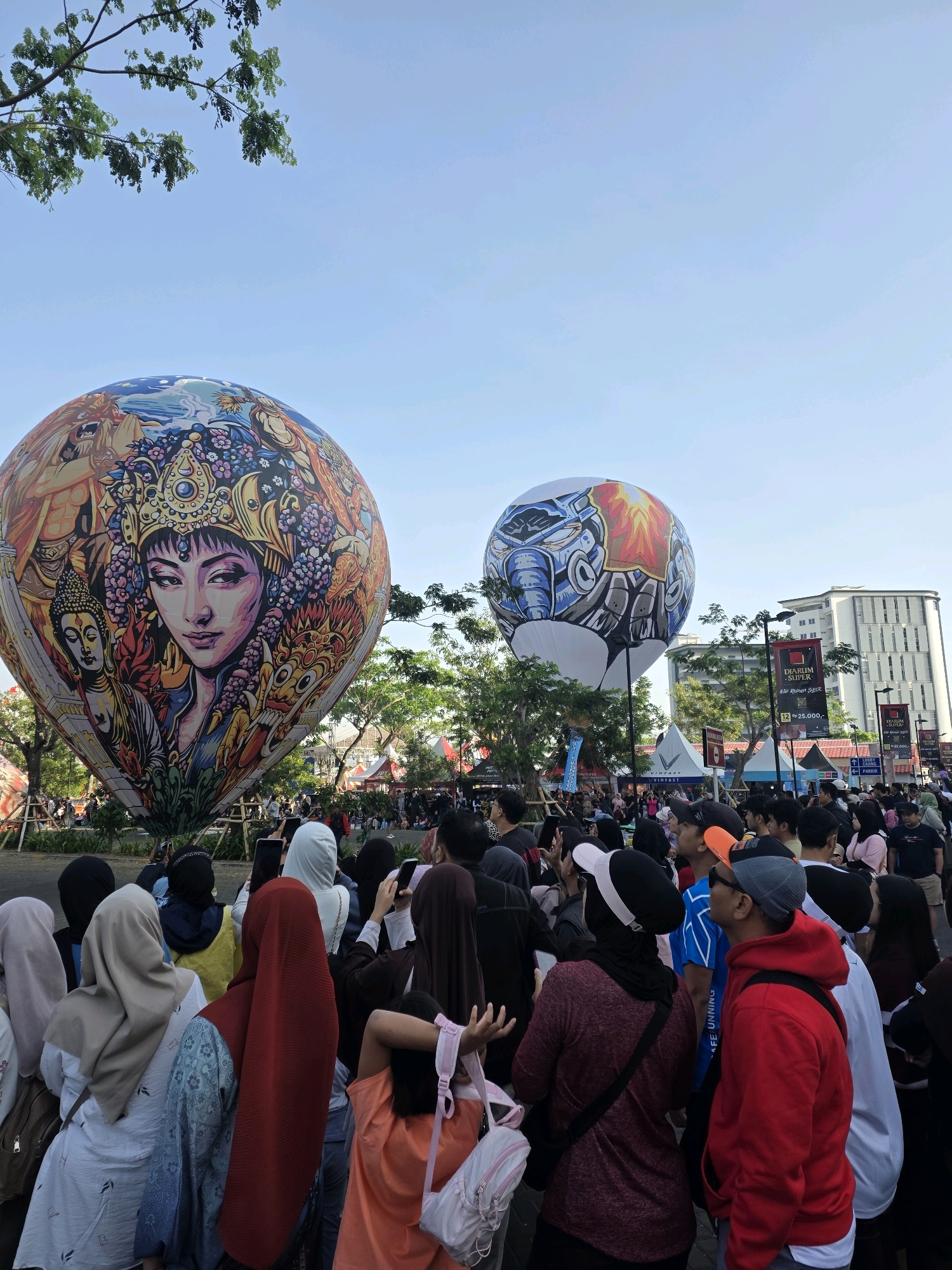 The image depicts an outdoor event with two large hot air balloons as the central subjects, displayed prominently in the foreground. The balloon on the left is adorned with a vibrant mural of a woman's face with intricate patterns and figures surrounding it. The balloon on the right features a graphic design resembling a robot or mechanical creature with fiery accents. Both balloons are tethered and appear to be part of a festival or exhibition.
A large crowd of people is gathered, observing the balloons and engaging in various activities. Many are holding up mobile phones to take pictures or videos. The crowd is diverse, with individuals of various ages and attire, many wearing casual clothing, some with headscarves. The people are densely packed, indicating a popular event.
The environment suggests an urban setting, possibly a park or open public space. In the background, several tents or vendor stalls are visible, along with trees, streetlights, and a tall modern building. Promotional banners are also present, with one clearly displaying "DIARUM SUPER" and a price. The sky is clear and blue, indicating daytime. The overall atmosphere seems lively and festive.
Based on the presence of promotional banners featuring brands like "DIARUM SUPER," and the general urban setting typical of such events, this scene is likely situated in a public space within the city of Bandung, Indonesia, known for hosting various festivals and public gatherings.