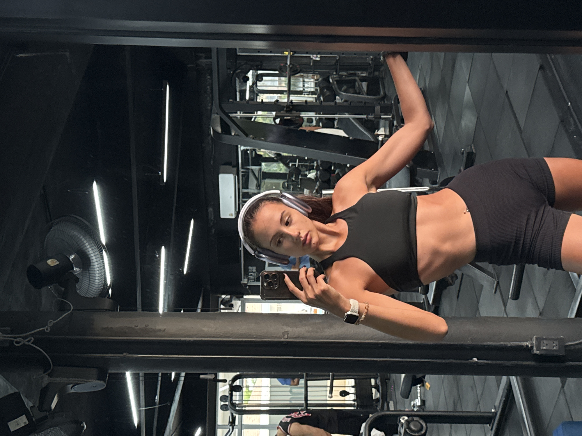 The image shows a person with light brown hair wearing a black sports bra and black shorts, posing in front of a mirror at a gym. The person is holding a phone and wearing headphones and a watch. Gym equipment is visible in the background. A fan is also present. Based on location data, this scene is in Kathu, Thailand.