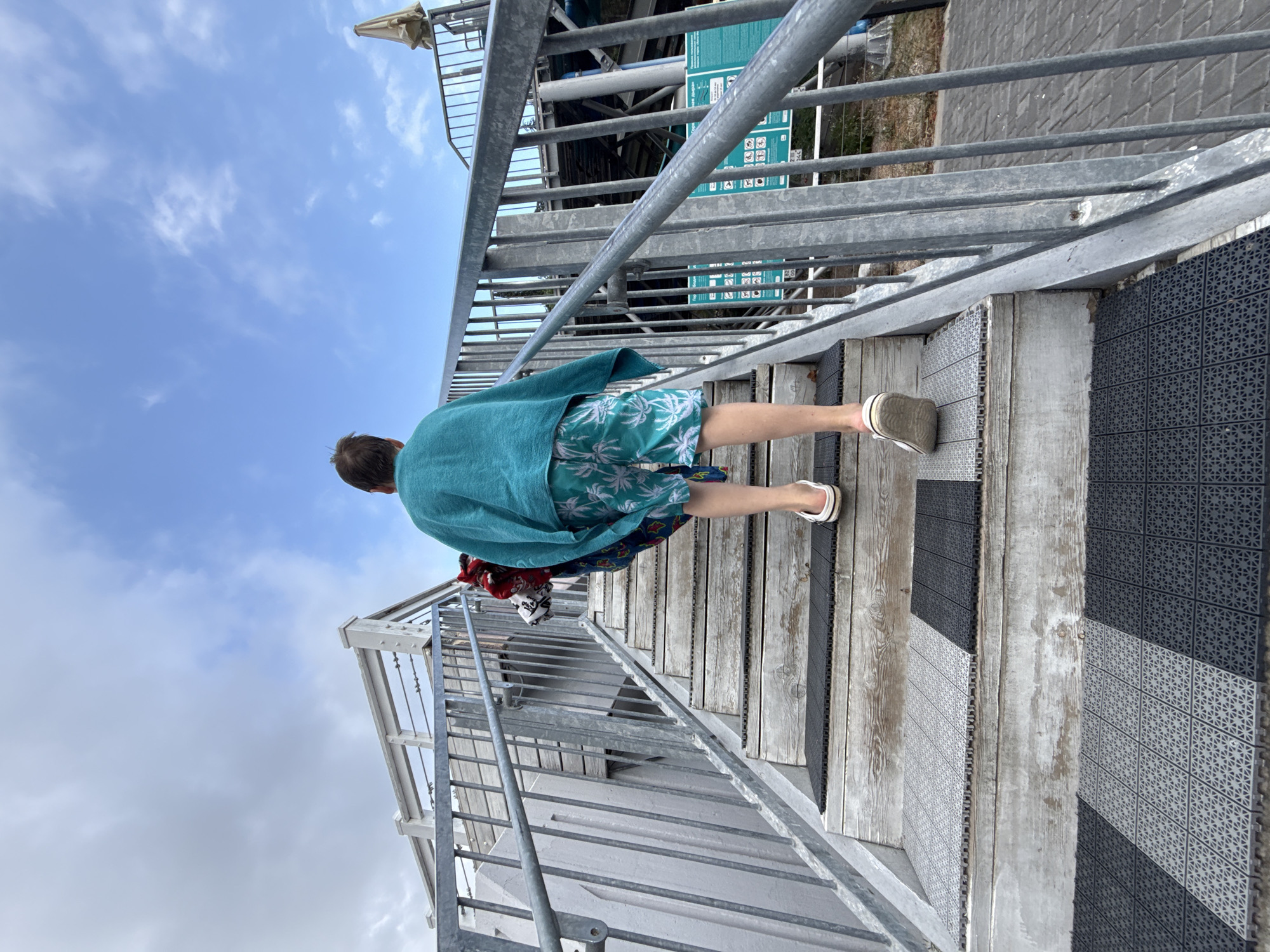 A young person, viewed from behind, walks up a set of outdoor stairs. They are wearing a teal towel draped over their shoulders and patterned swim shorts. The stairs are constructed of wood with metal railings on both sides. The steps are covered with textured mats, alternating in color between black and grey. The sky above is blue with scattered white clouds. In the background, part of a building with a white railing and an umbrella are visible. This scene appears to be located in a recreational area, possibly near a swimming pool or beach facility, in the city of Yalta, Ukraine.