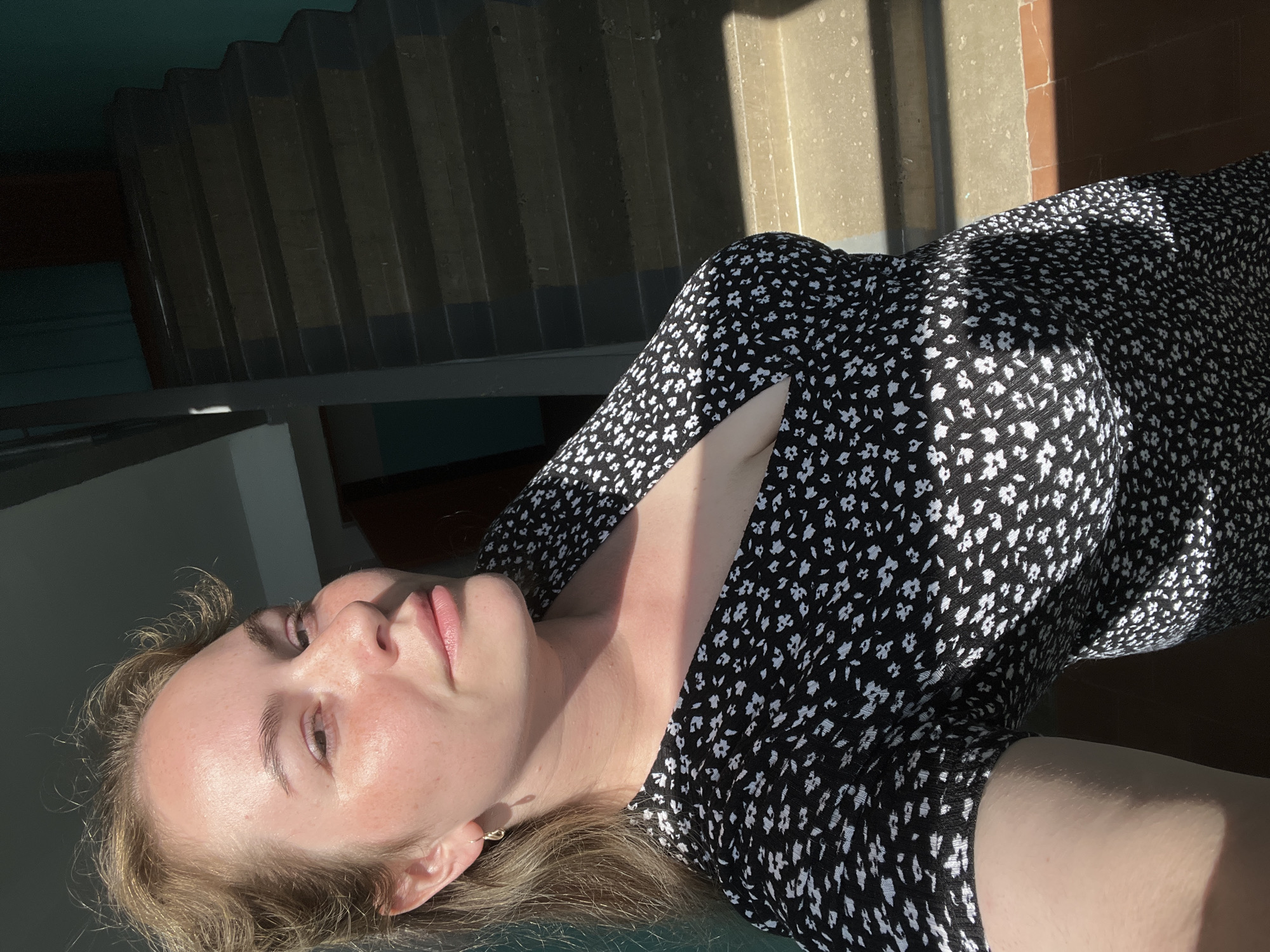 A fair-skinned woman with blonde hair is pictured from a low angle, lying down, wearing a black dress with small white floral print. She is looking upwards, slightly towards the left. A bright, angled sunlight illuminates her face and chest. In the background, stairs and a wall can be seen, cast in shadow and light. The location is Chaikovsky, Russia.