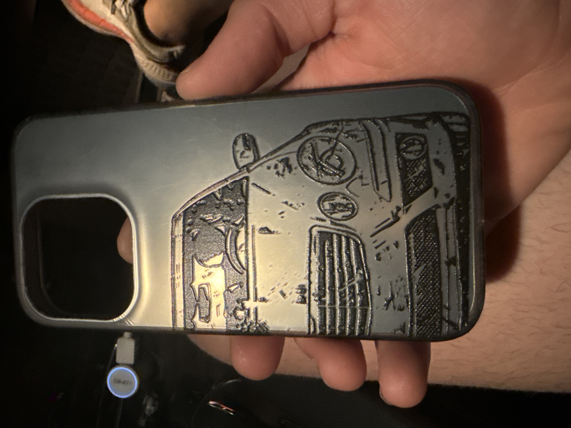 An embossed image of a car's front end is etched onto a silver-colored phone case, held by a person's hands. The case is rectangular with a large cutout at the top for the phone's camera. The car depicted is a dark sedan with a prominent grille, headlights, and side mirror. The image is detailed, showing the car's contours and features. A white USB cable with a blue indicator light is visible to the left of the phone case. In the background, a person's leg and foot, wearing a white and orange shoe, are visible to the right. The environment appears to be dimly lit, possibly indoors or at night. The image does not provide any information about the specific location in the city of Stara Zagora, Bulgaria.