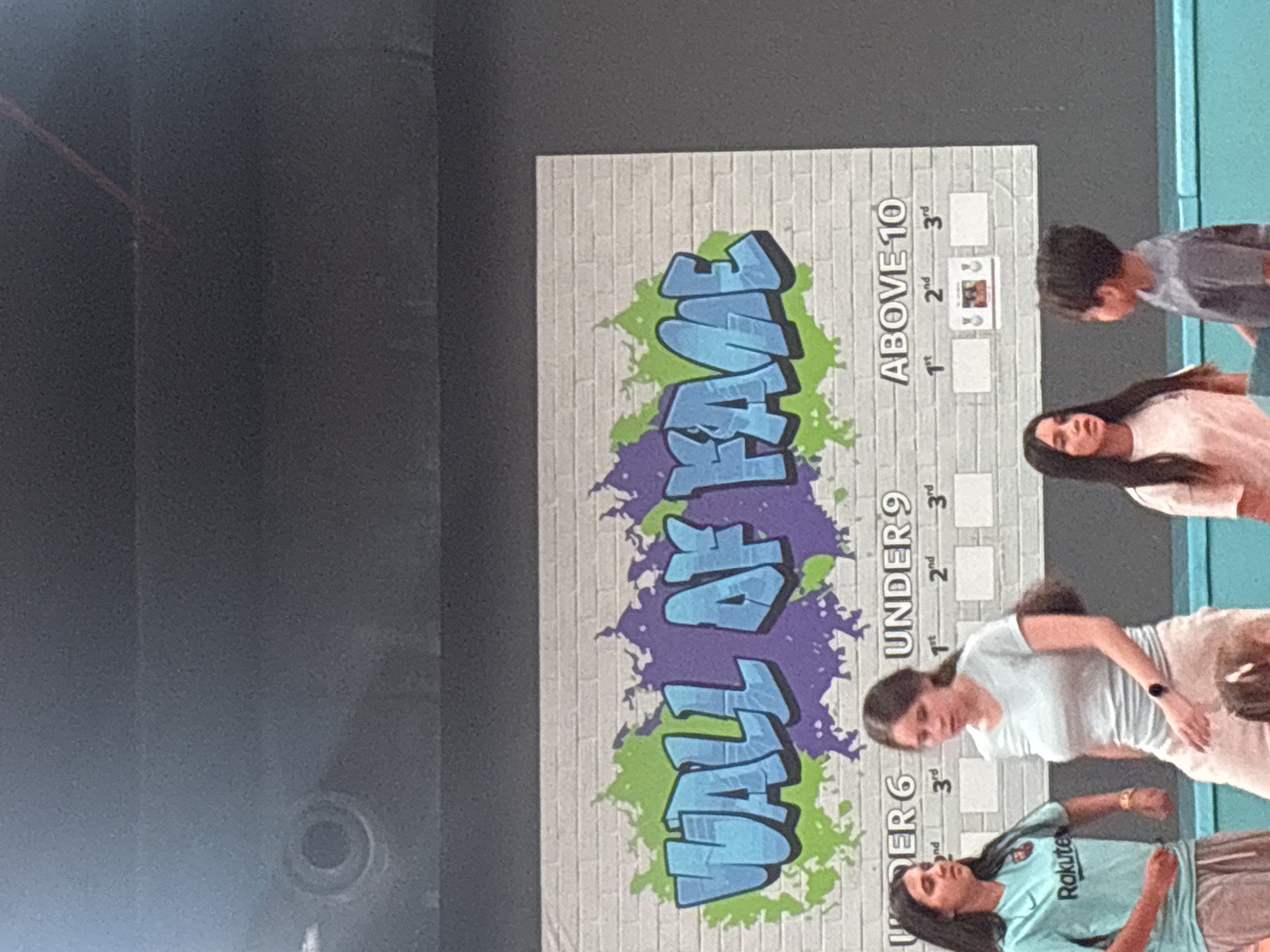 This image captures a group of individuals, predominantly females of various ages, in what appears to be an indoor recreational facility, possibly a trampoline park or similar venue. Dominating the background is a large banner with graffiti-style lettering that reads "WALL OF FAME." Below this, a chart is organized into age categories: "UNDER 6," "UNDER 9," and "ABOVE 10," each with sub-sections for "1st," "2nd," and "3rd." There are blank squares for entries, and in the "ABOVE 10" category, a small framed picture is visible in the "2nd" place square.
Two young women stand in the foreground on the left. The woman on the far left is wearing a turquoise t-shirt with a "Rakuten" logo and grey sweatpants. She is looking to her left. Beside her, a woman in a light blue t-shirt and light-colored pants is looking downwards and slightly to her right. In the center right of the image, a young girl in a pink shirt is looking to her right, and a young boy in a grey t-shirt is facing away from the camera, looking towards the girl. The overall environment suggests a place for competitive activities or achievements. The presence of trampoline-like blue padding is visible at the bottom of the frame. The lighting suggests an indoor setting, possibly during the day or with artificial lighting. There is no specific indication of the location being in the city of Dubai, United Arab Emirates.