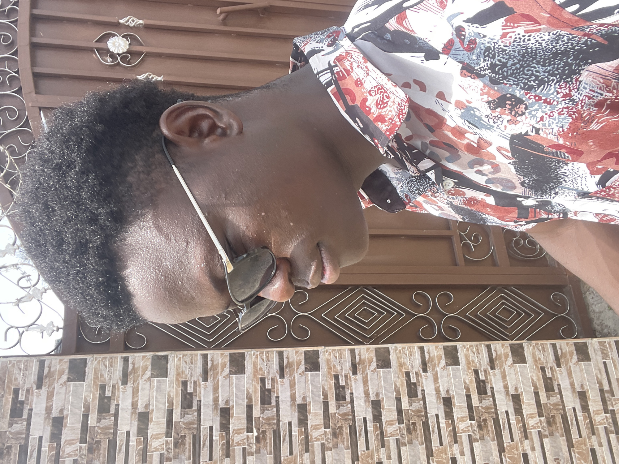 A medium shot shows a Black male with short, curly hair and brown skin wearing a colorful patterned shirt and sunglasses. He is positioned against a background of a brown metal gate with decorative geometric and floral patterns, which rests above a section of tiled wall with a vertical stacked pattern. The image appears to have been taken outdoors in Badagry, Nigeria.