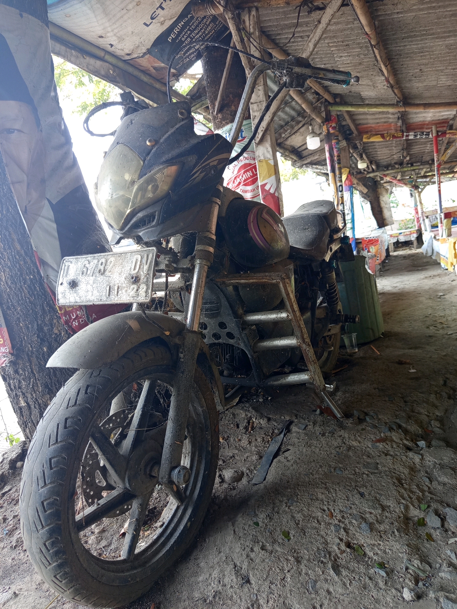 An abandoned motorcycle, identified by license plate D 6782 DV and an expiry date of 11-22, is parked outdoors under a makeshift shelter constructed from corrugated metal and bamboo. The motorcycle is covered in dust and cobwebs, indicating a prolonged period of disuse. The scene appears to be a roadside or outdoor market setting, possibly in Karawang, Indonesia, suggested by the presence of similar makeshift stalls and signage in the background. The ground is a mix of dirt and gravel, with scattered debris and leaves. The overall environment is weathered and appears somewhat neglected. There are no people visible in the image.